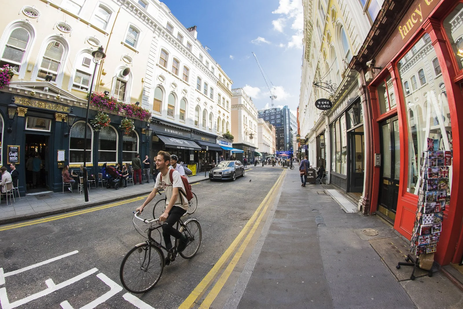A city street scene in London with a man riding a bicycle in the foreground, surrounded by shops, cafes, and buildings with colorful facades, under a blue sky with a few clouds.