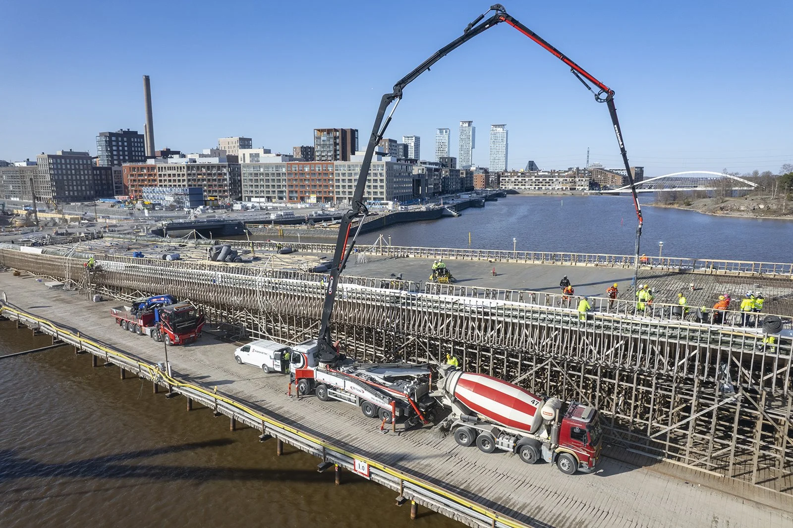 Construction workers are building a bridge over a body of water with concrete mixer trucks and large cranes, with city buildings in the background during daytime.