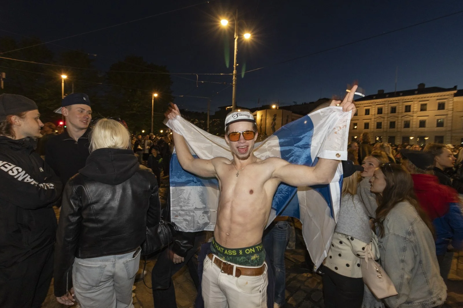 A shirtless man holding a Finnish flag and wearing sunglasses, surrounded by a crowd of people celebrating Finland's hockey world cup victory.