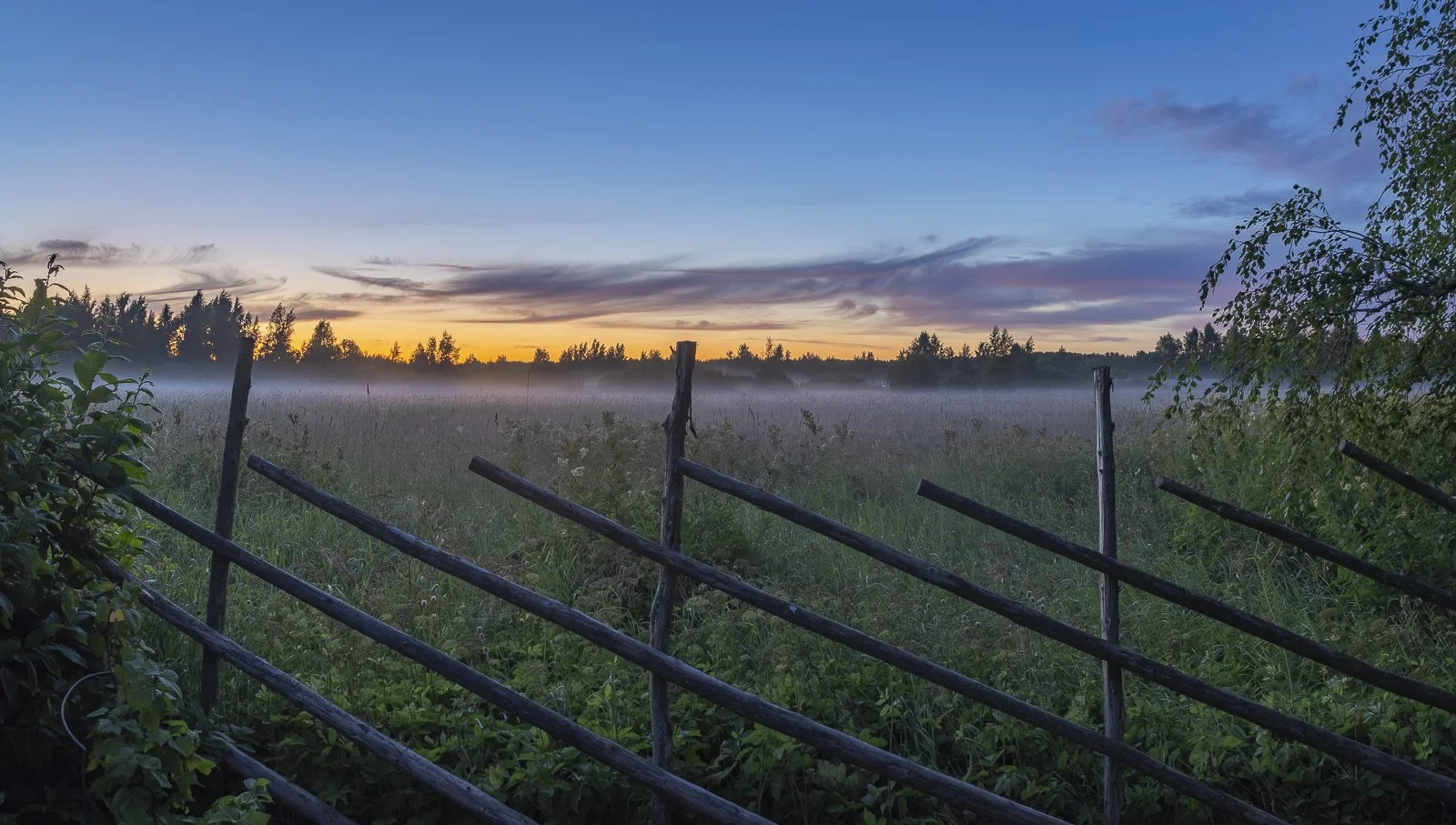 A rural landscape at dawn with a wooden fence in the foreground, a grassy field, and a line of trees under a sky with sunrise colors and some clouds.