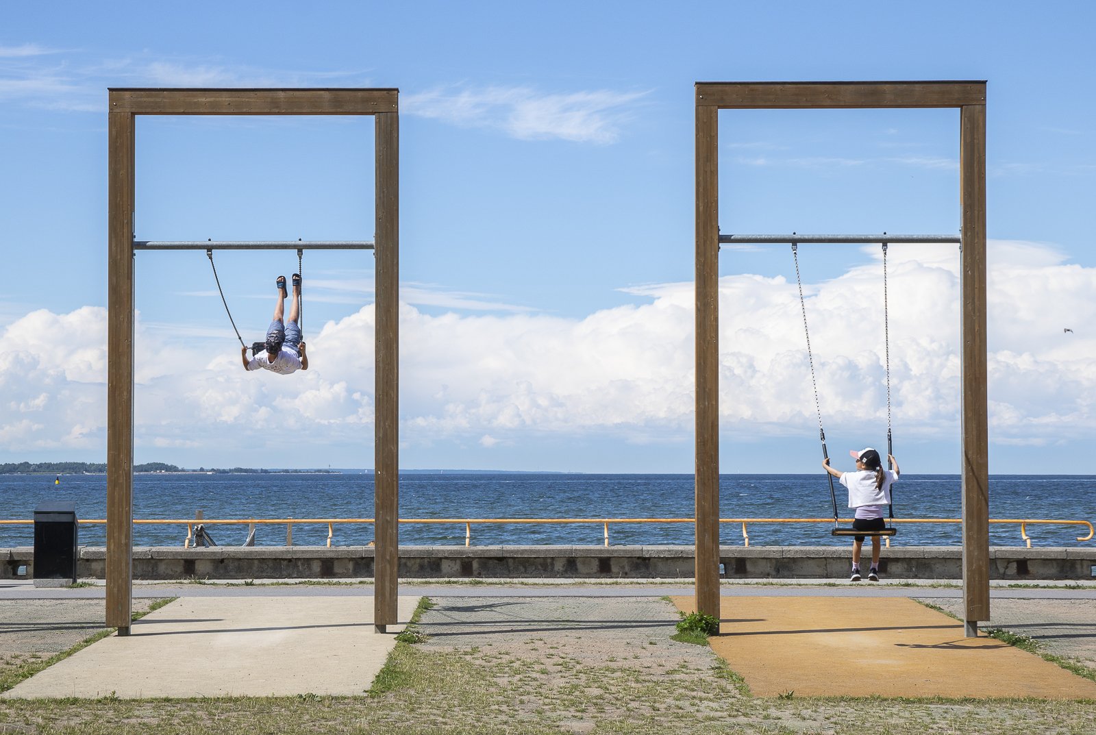 Children playing on swings near the water with a cloudy sky in the background.