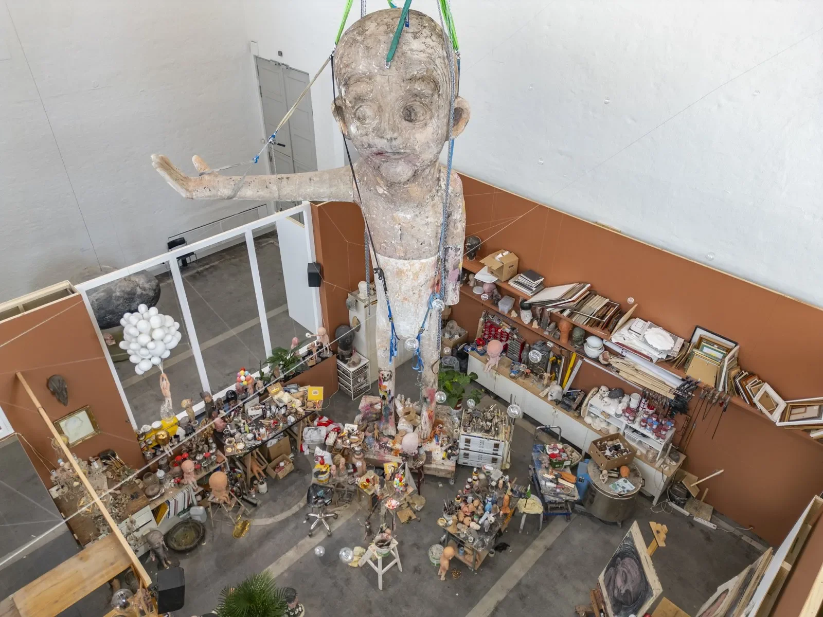 An art studio with a large sculpture of a child's head and upper body suspended from the ceiling, surrounded by shelves and tables filled with art supplies, sculptures, paintings, and miscellaneous objects.