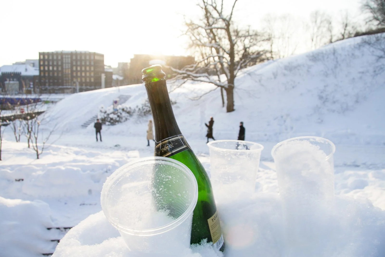 A champagne bottle and several empty plastic cups in the snow with a snowy park and people walking in the background at sunrise in Tallinn, Estonia.