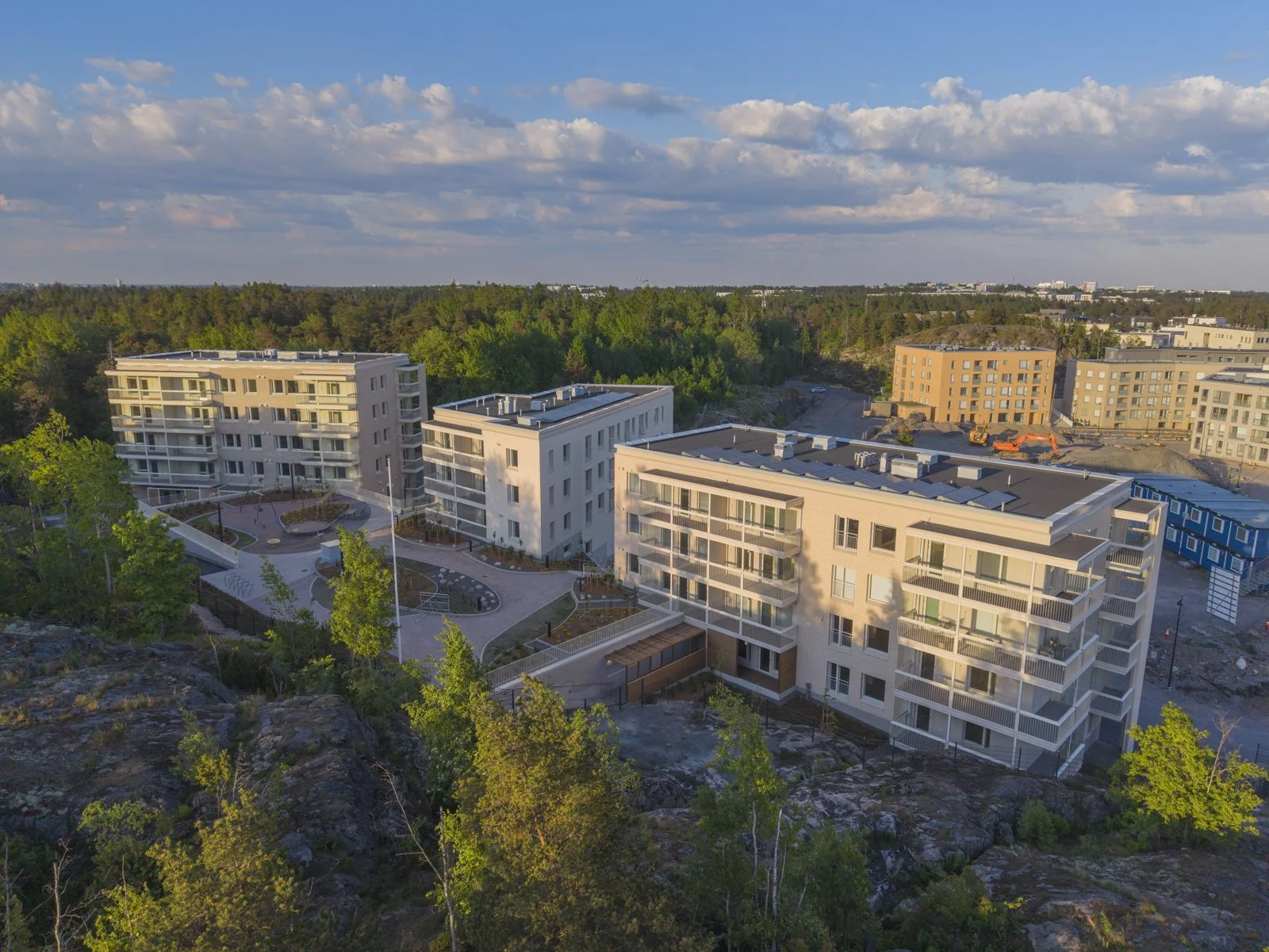 Aerial view of newly constructed residential buildings with balconies, a landscaped courtyard, and construction equipment on a partly cloudy day, with a forest in the background in Helsinki, Finland.