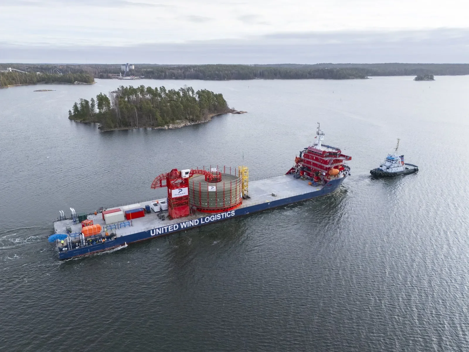 A large blue cargo ship labeled 'United Wind Logistics' is being guided by a small tugboat on a body of water. The cargo ship is carrying equipment with a red and yellow structure on top, and nearby are storage containers and safety equipment. Land w