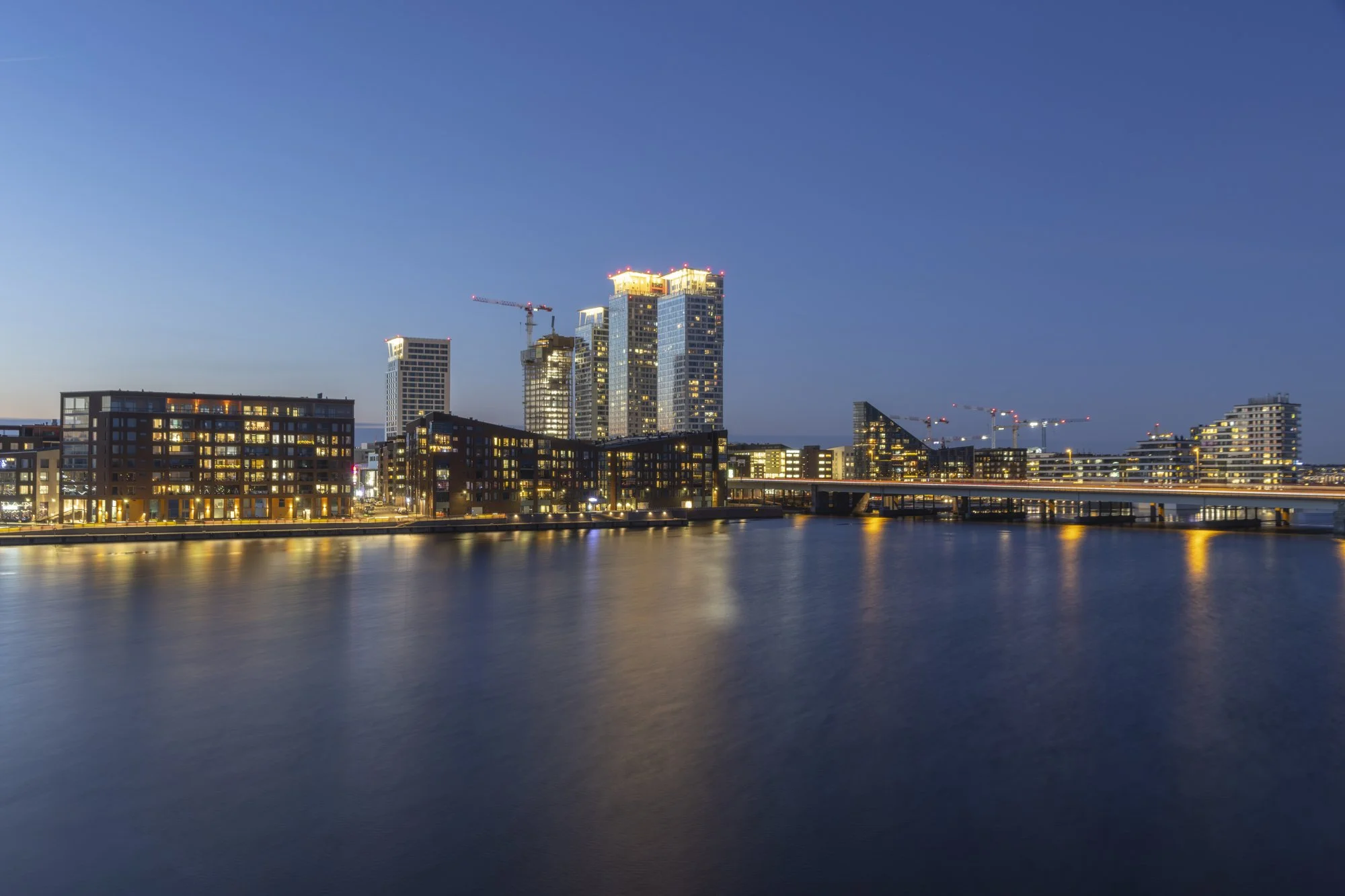City skyline in Kalasatama district in Helsinki, Finland during twilight with illuminated modern high-rise buildings, including some under construction, alongside a body of water reflecting the city lights.