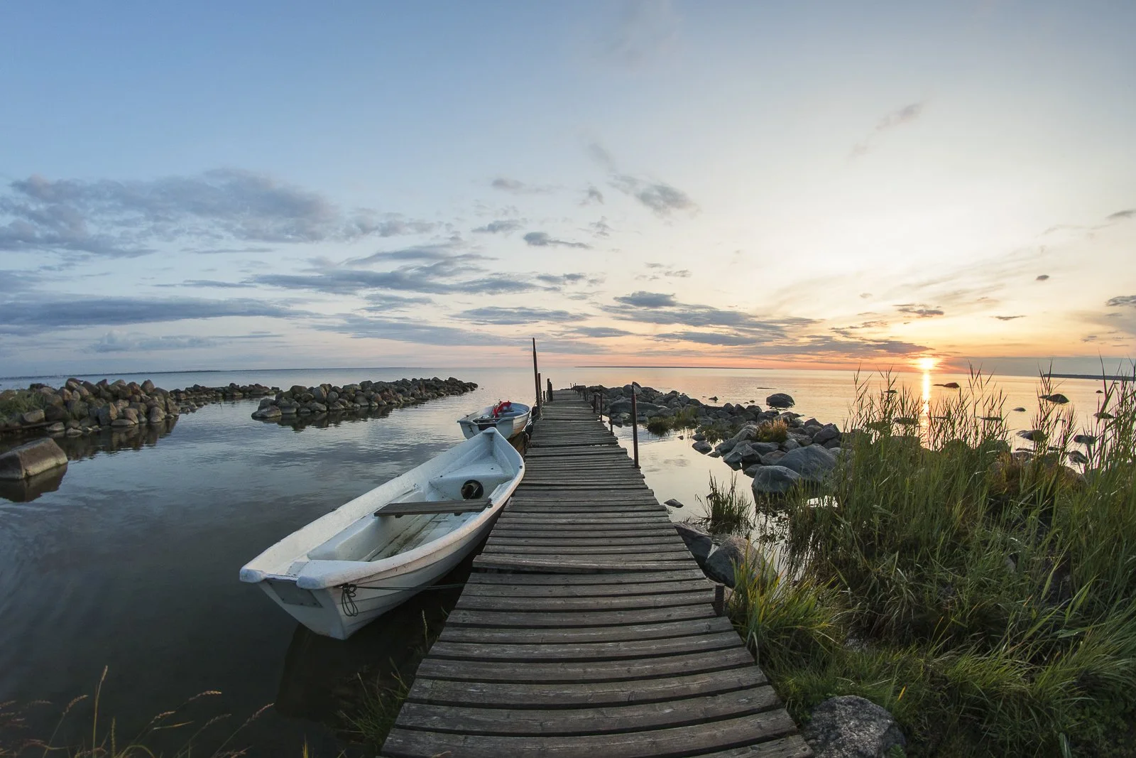 A wooden dock extends into a calm body of water at sunset, with small boats moored along the sides and rocks and grass nearby, under a partly cloudy sky.