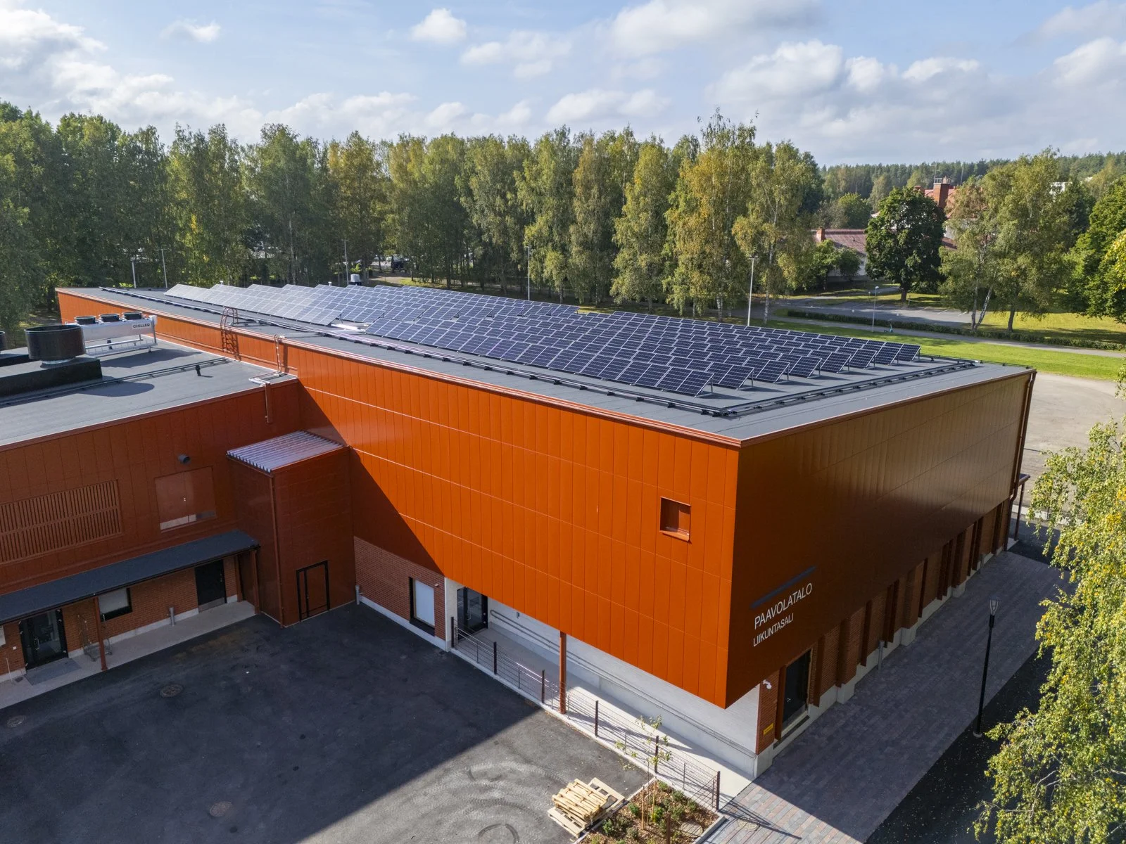 Aerial view of a modern building with solar panels on the roof, surrounded by trees and a parking lot, under a partly cloudy sky.