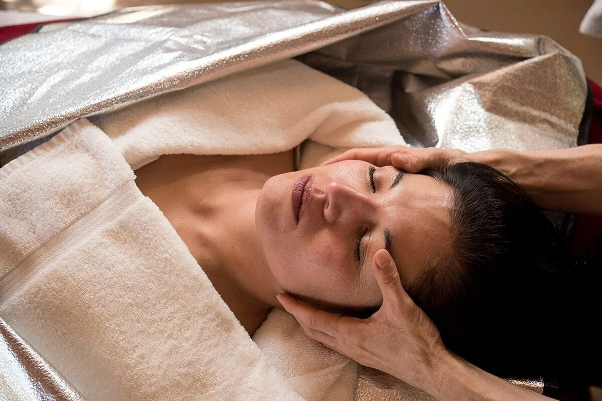 A woman receiving a massage, lying on her back with eyes closed, while a therapist gently holds her head.