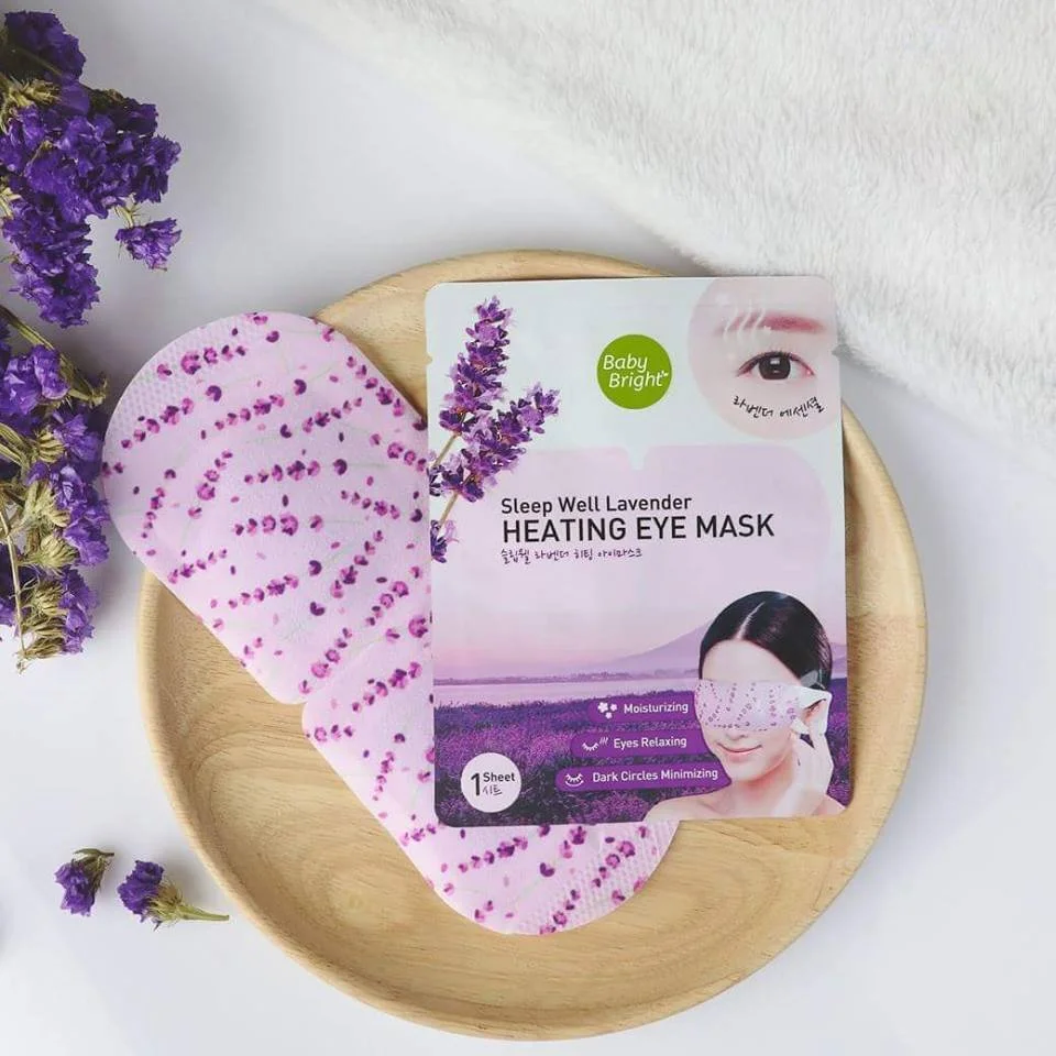 Packaging of Sleep Well Lavender Heating Eye Mask with purple lavender flowers on the side, placed on a light wooden plate, with a white textured cloth in the background.
