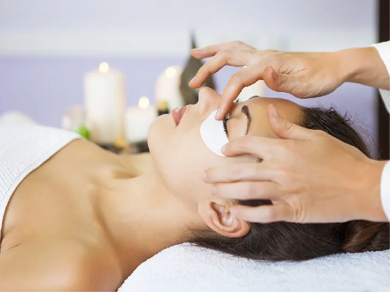 Woman receiving facial treatment at spa or skincare clinic, lying on a treatment bed with eyes closed, with hands of a professional applying skincare product.
