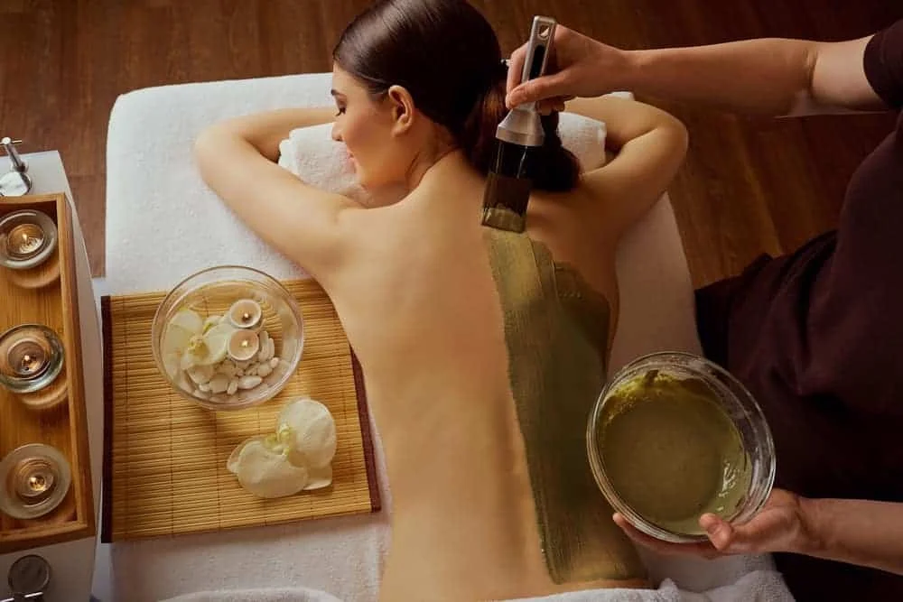 A woman receives a back massage in a spa, with a therapist applying a green herbal body mask.