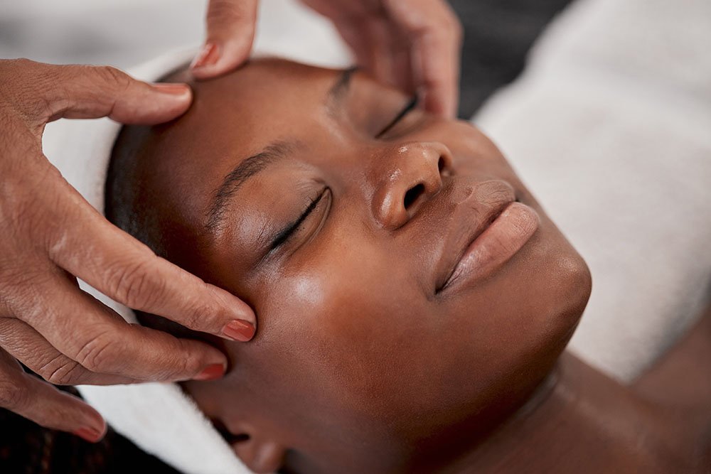 A person receiving a facial massage with their eyes closed, lying down, with hands gently massaging temples.
