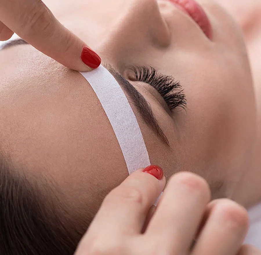 A person with long eyelashes has a white strip being placed on their eyelid by another person with red painted nails.