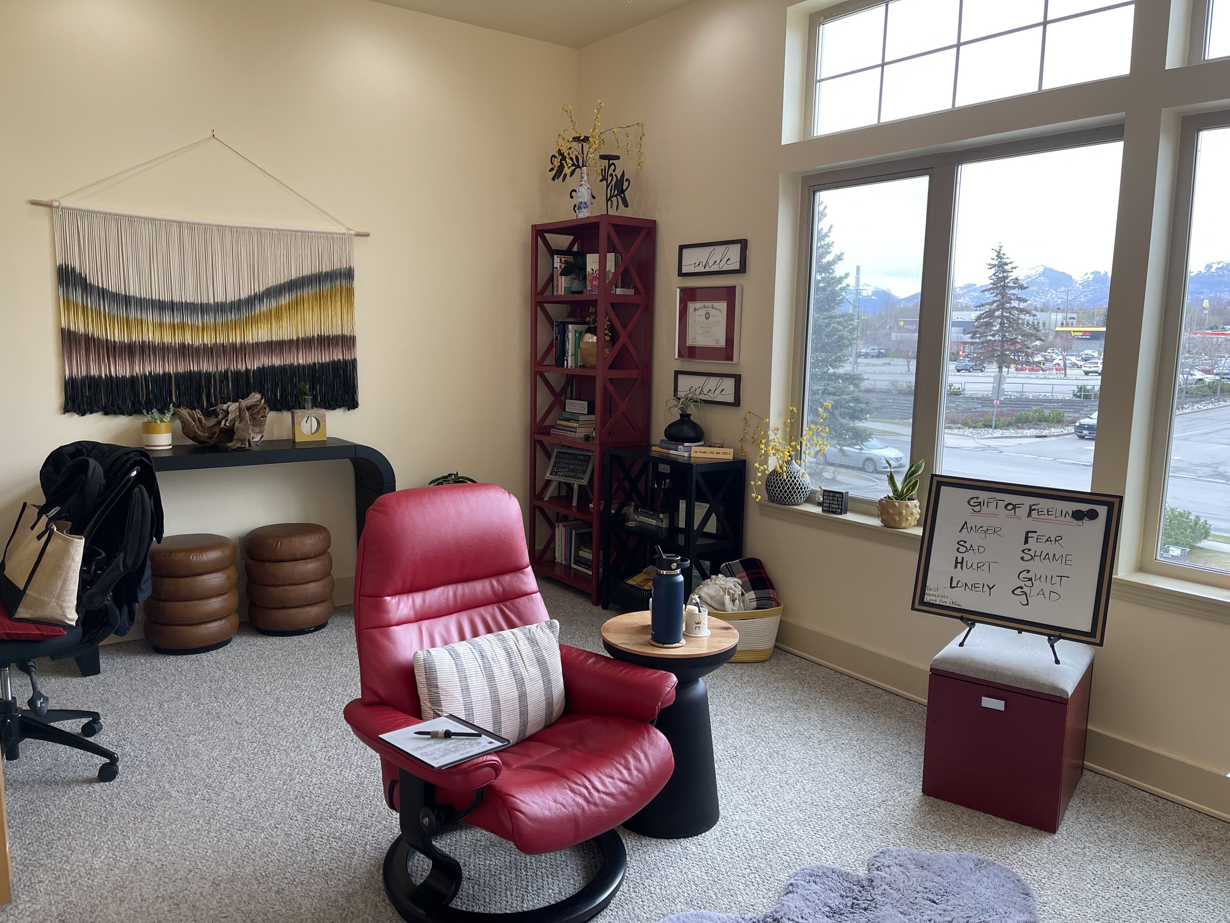 Indoor office space with a red desk chair, a round side table with water bottle and tissues, a whiteboard on a red box with feelings written on it, large windows showing parking lot and mountains, and various decorative items.