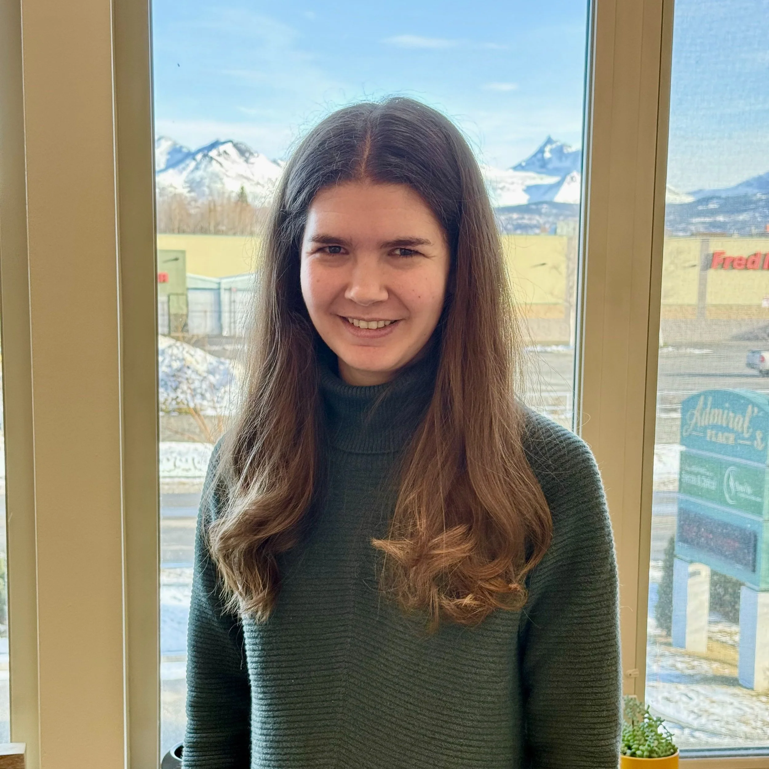 Elizabeth, wearing a dark green turtleneck sweater, standing indoors near a window with a mountain view in the background.