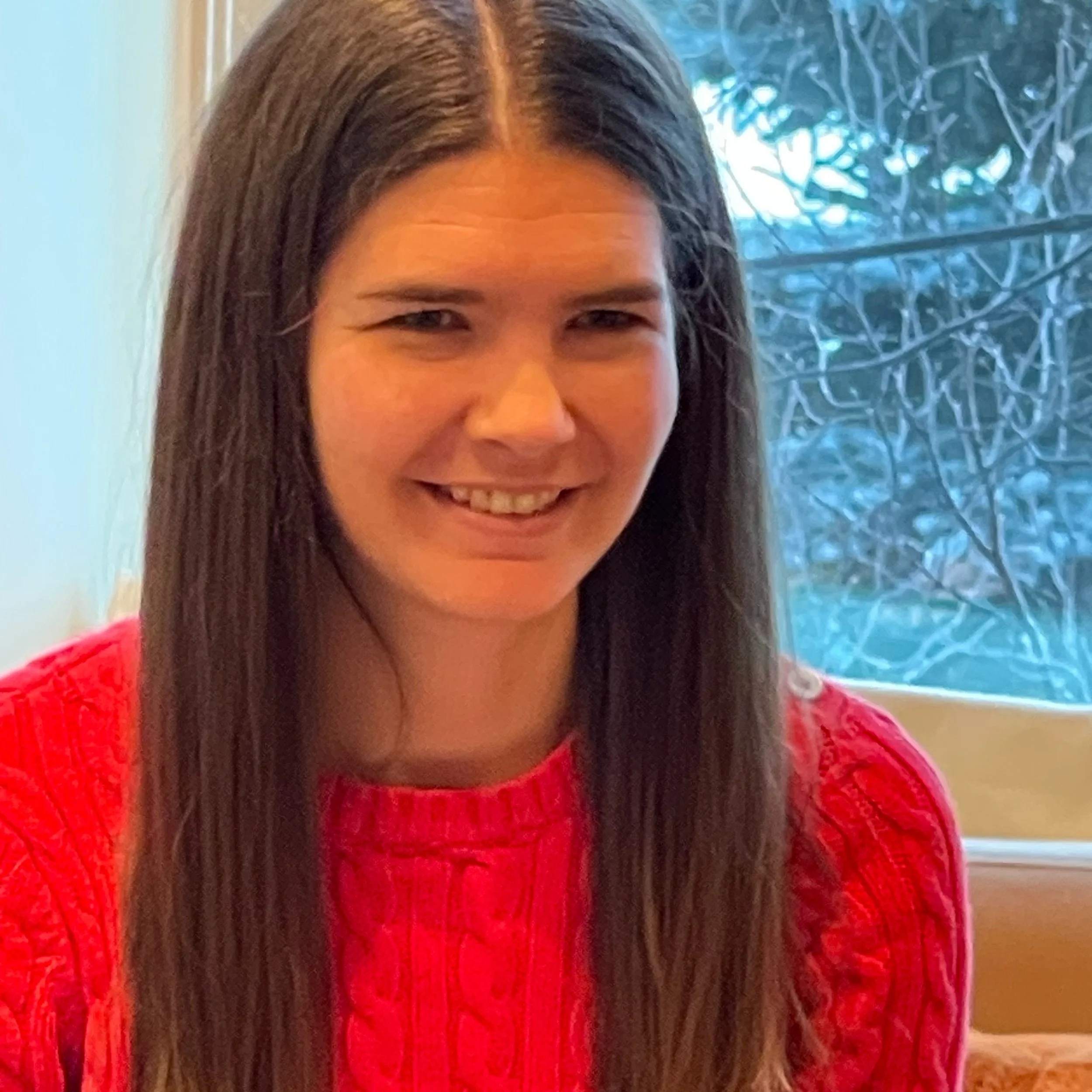 A woman with long brown hair wearing a red sweater, smiling indoors near a window with trees outside.