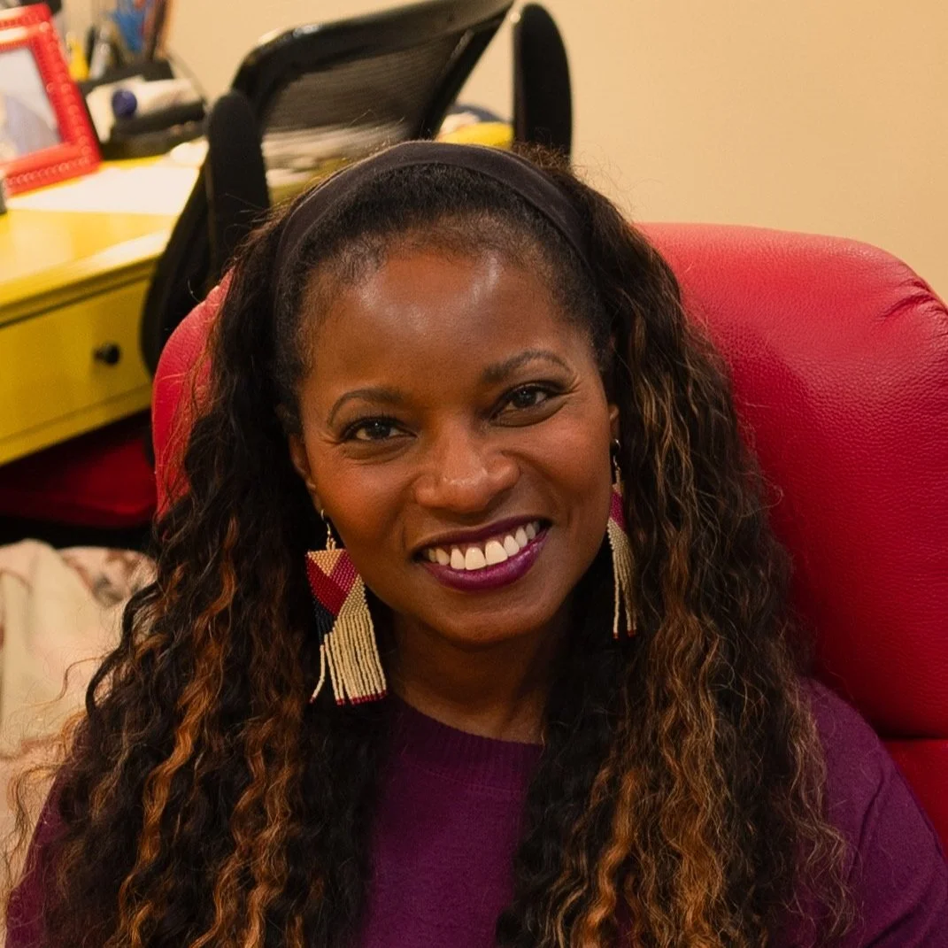 A woman with long wavy hair sitting on a red chair, smiling, wearing a purple top and colorful earrings.