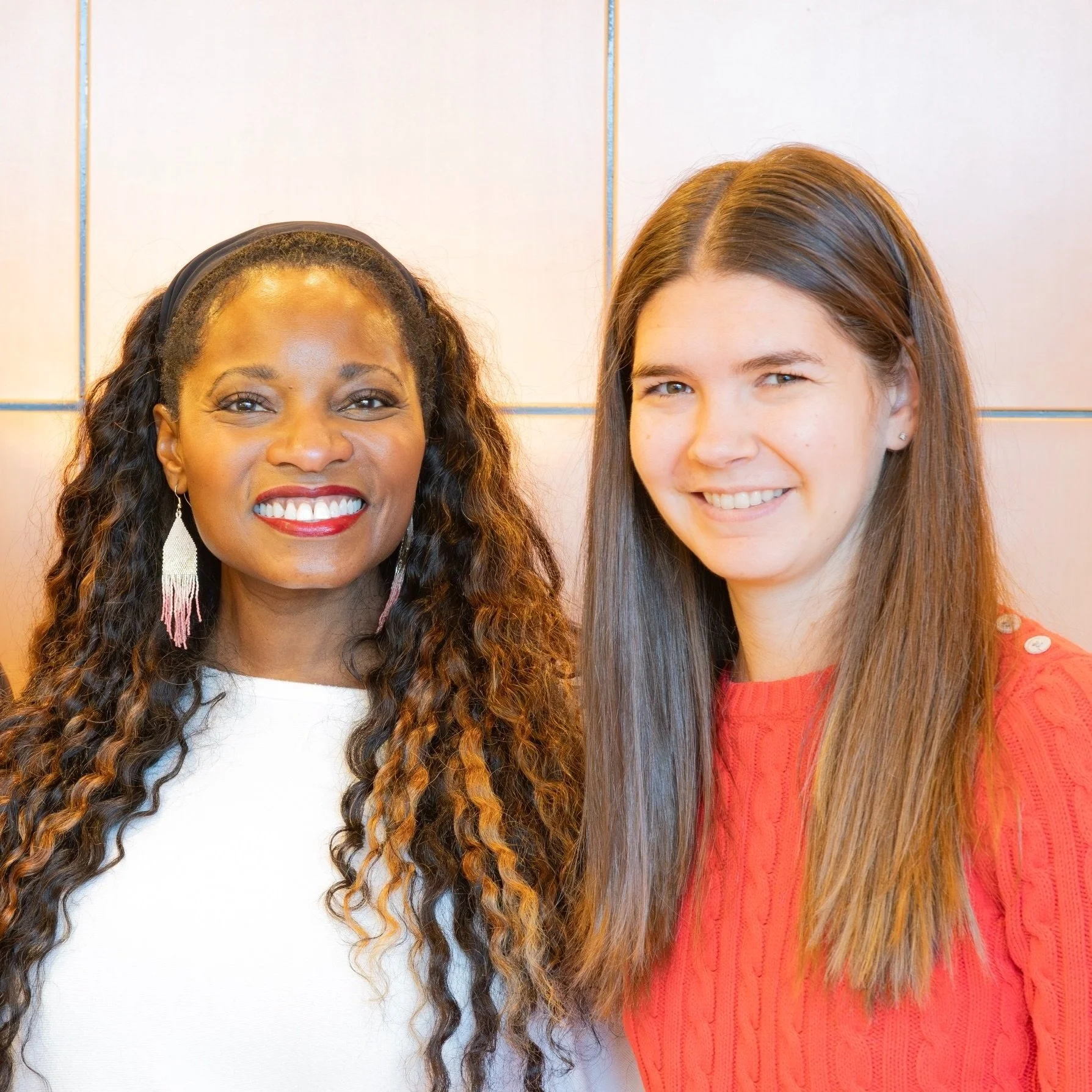 Two women smiling side by side in front of a light-colored wall.