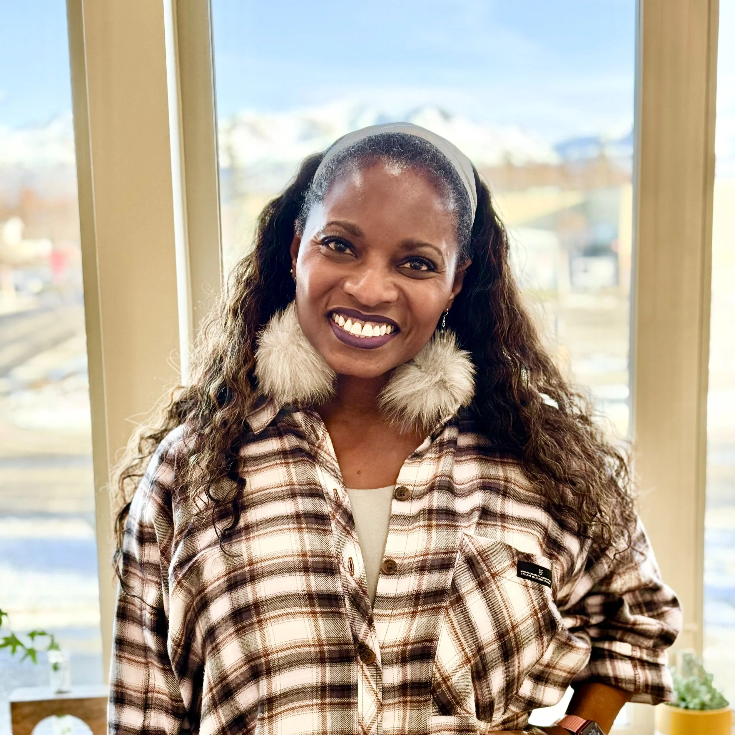 A woman with dark curly hair wearing a plaid shirt with faux fur collar accessories standing in front of a window with a scenic mountain view.