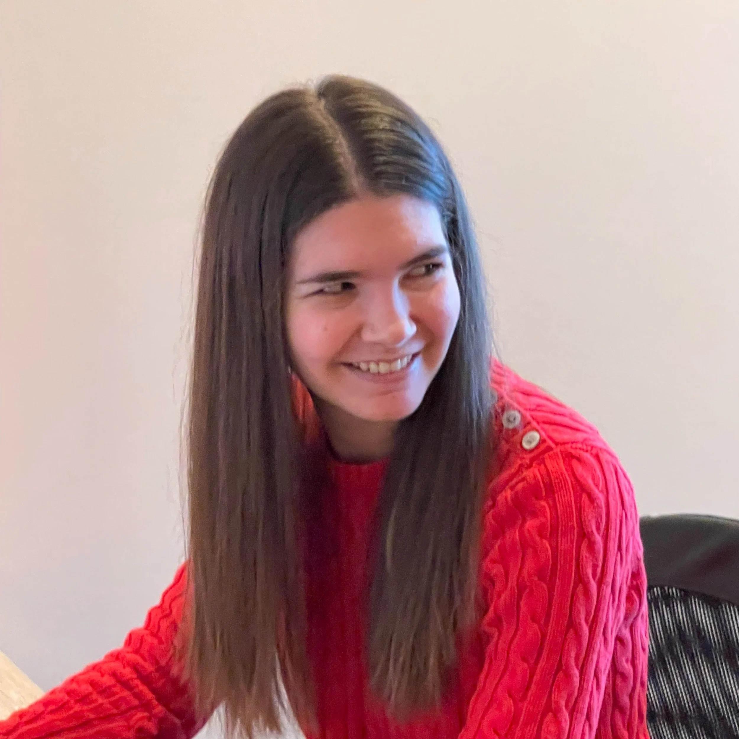 A young woman with long brown hair wearing a red cable-knit sweater with buttons on the shoulder, sitting at a desk with a smile and looking to her right.