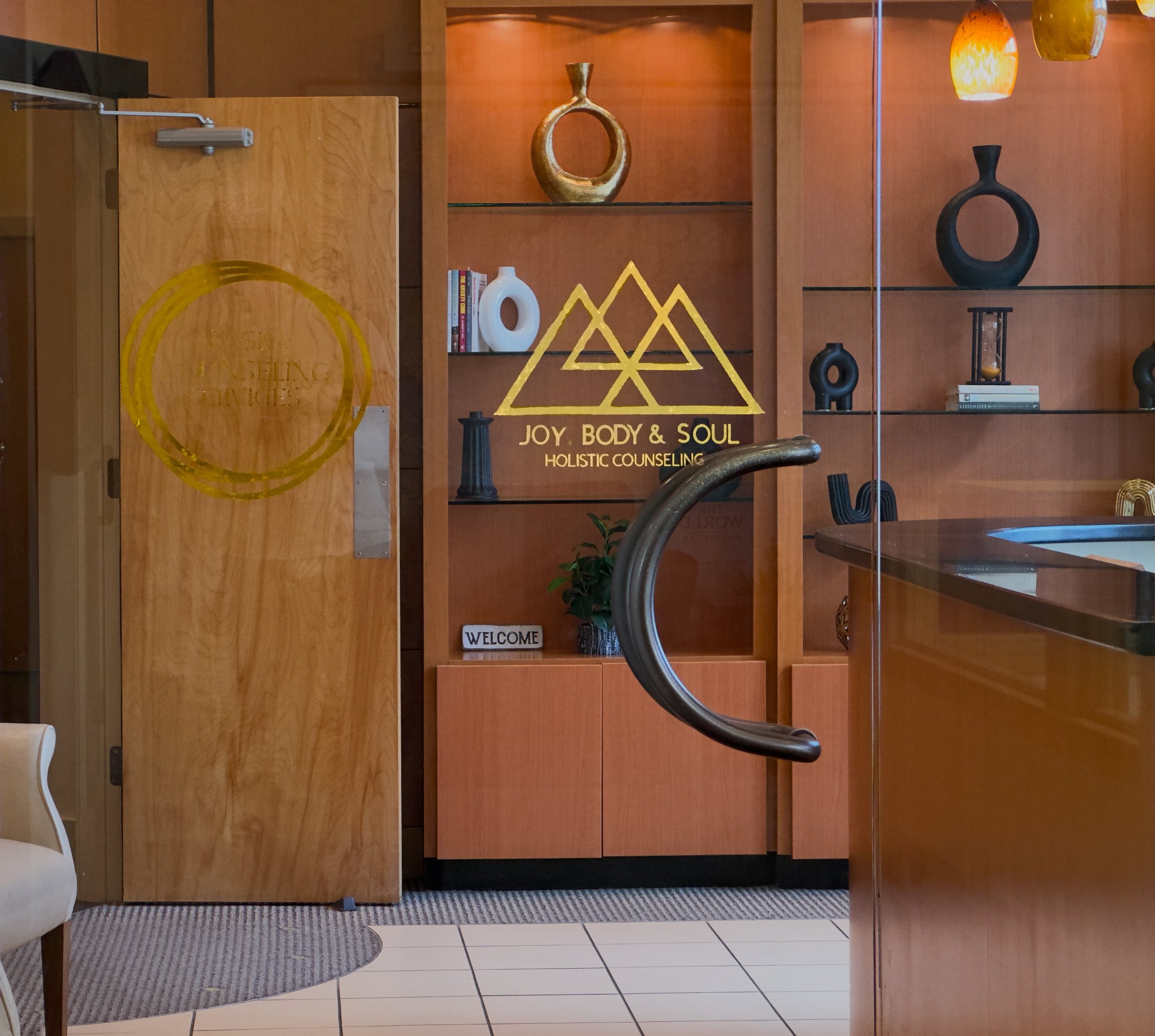 Interior of a holistic counseling office with wooden walls and shelves, decorative vases, and a welcoming sign. A wooden door with a gold painted logo is slightly open.