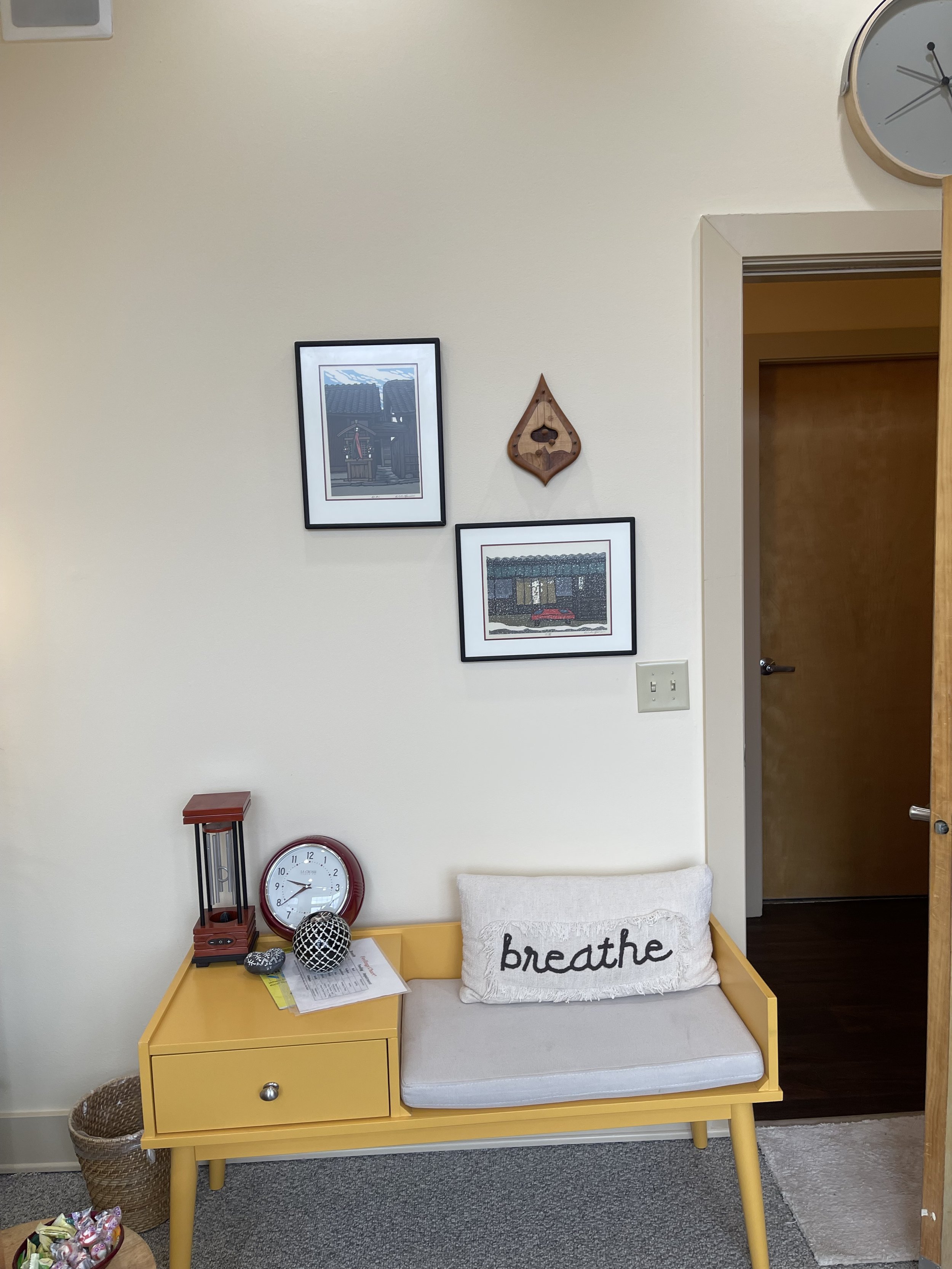 Corner with yellow bench, white pillow with 'breathe' text, clock, framed pictures, and wall art.