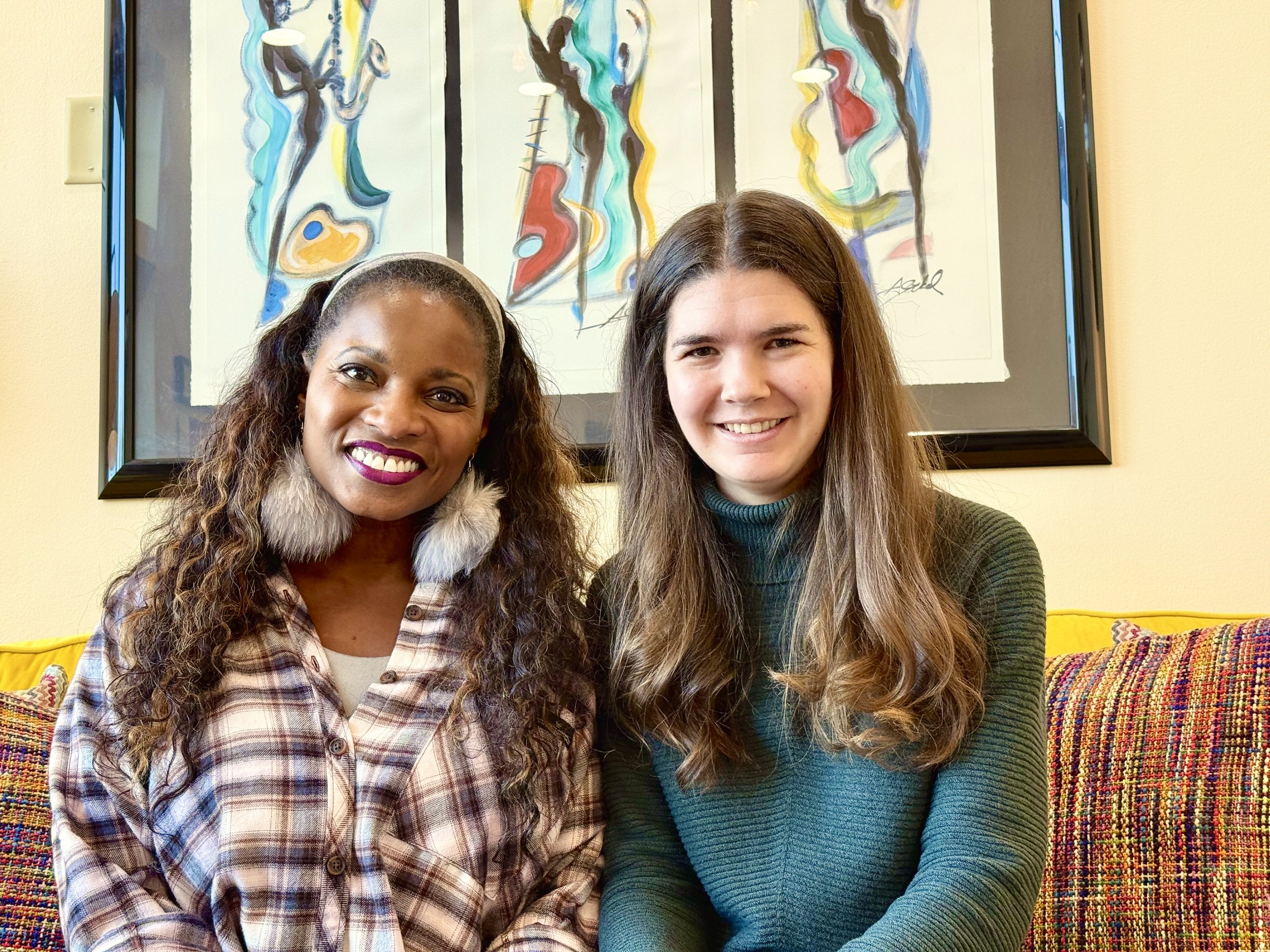 Two women sitting on a colorful couch in front of abstract artwork with vibrant colors and patterns, smiling at the camera.