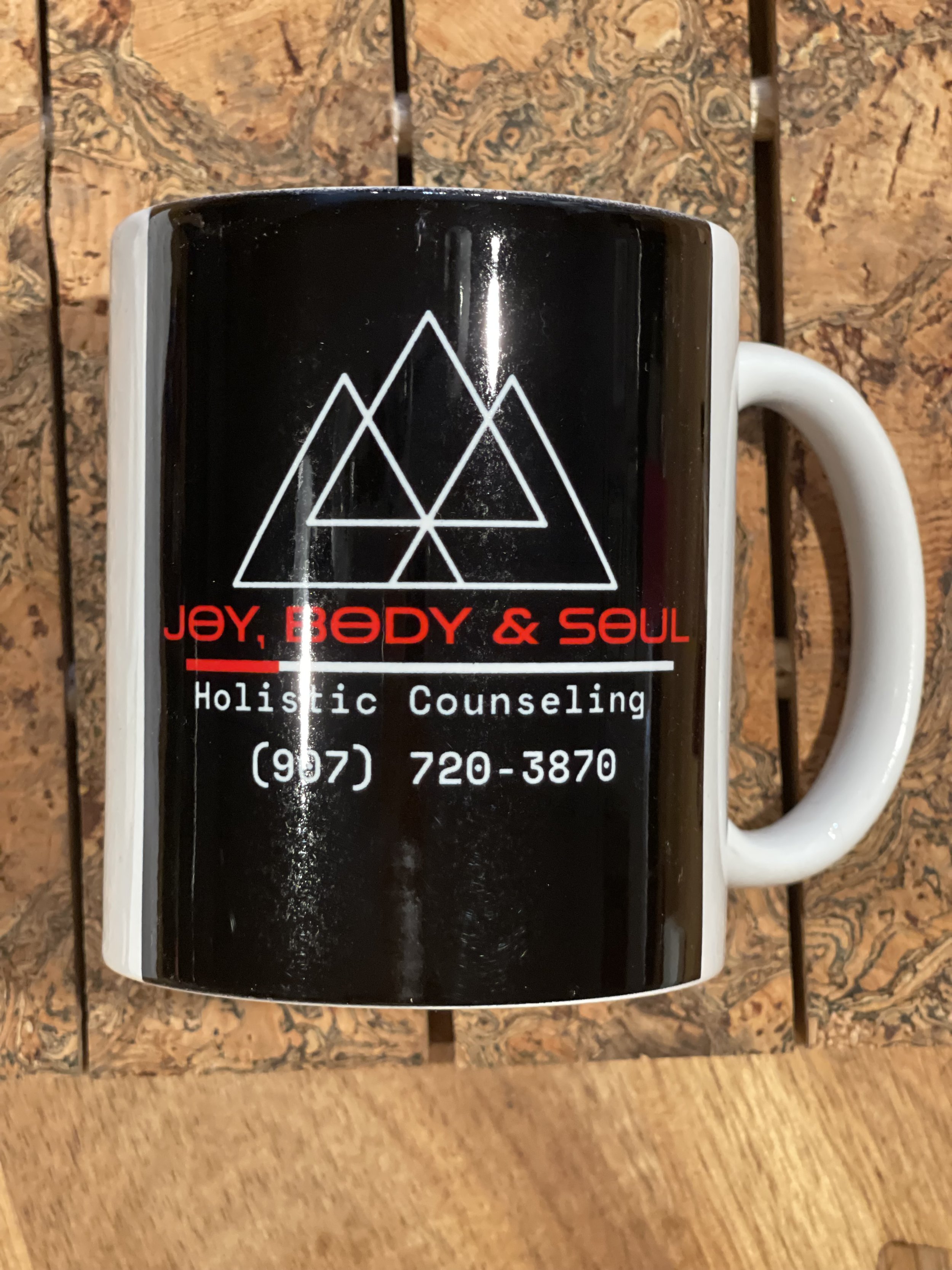 Black mug with white geometric design and red and white text promoting holistic counseling, resting on a wooden surface.