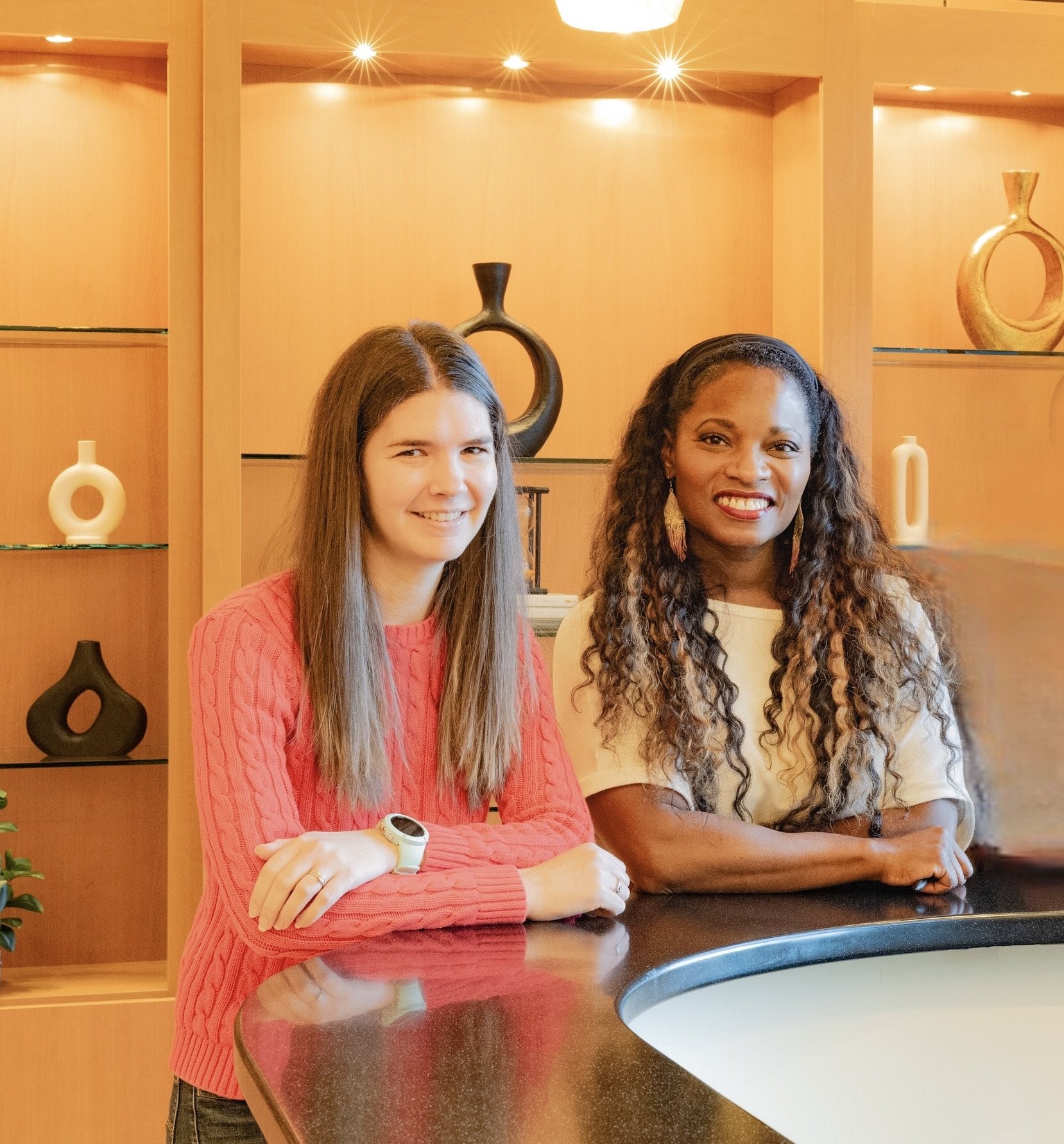 Two women, one with long straight hair wearing a pink sweater and the other with curly hair wearing a white top, smiling and leaning on a black countertop in a room with wooden shelves and decorative vases.
