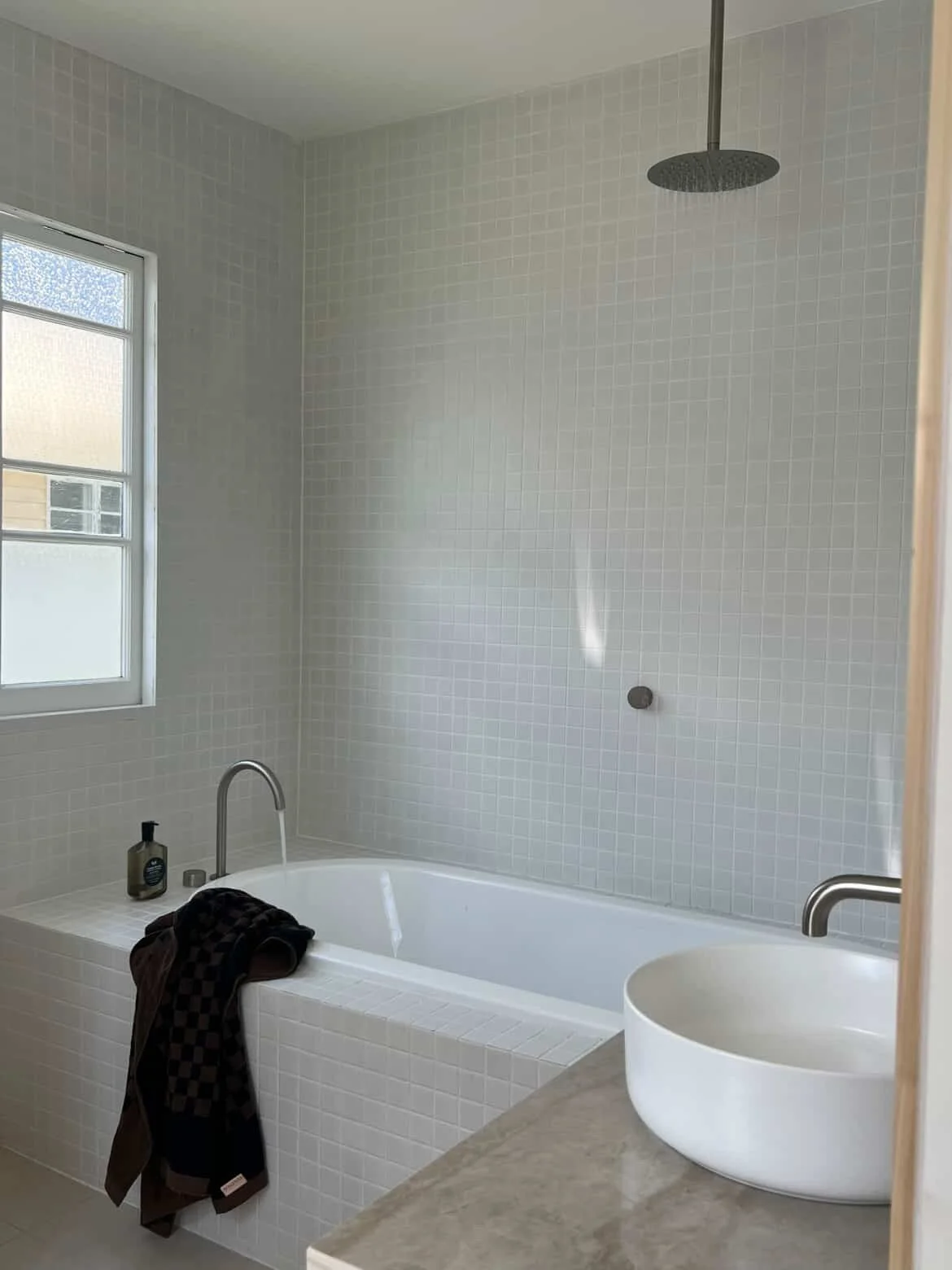 A bathroom with a bathtub, a small bowl sink on a countertop, and a window. The bathtub has black and brown towels hanging over the edge, with shampoo and soap bottles on the tub's edge. The showerhead is mounted on the white tiled wall, and natural 