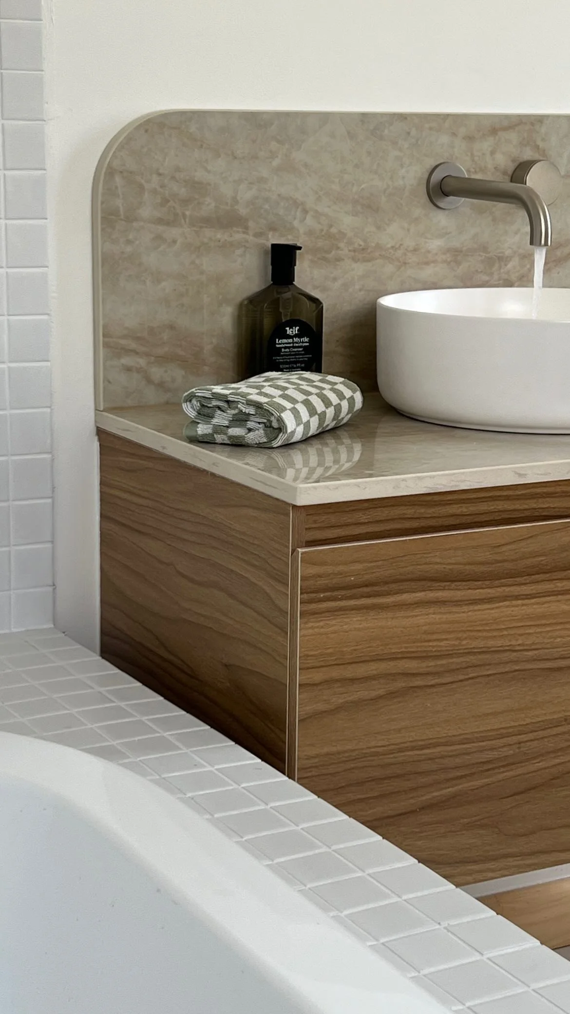 Bathroom renovation with marble stone countertop, warm cabinetry, with a small round vessel sink, a wall-mounted faucet, a bottle of body cleanser, and a rolled checkered towel. Sunshine Coast, Noosa, Brisbane.