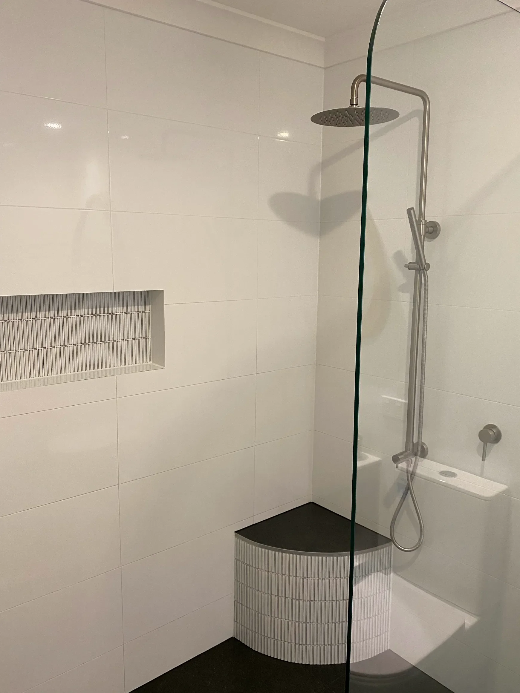 Modern shower area with a rain showerhead, a handheld showerhead with a hose, a small built-in shelf, and a curved step with white tile and a dark countertop in a bathroom.