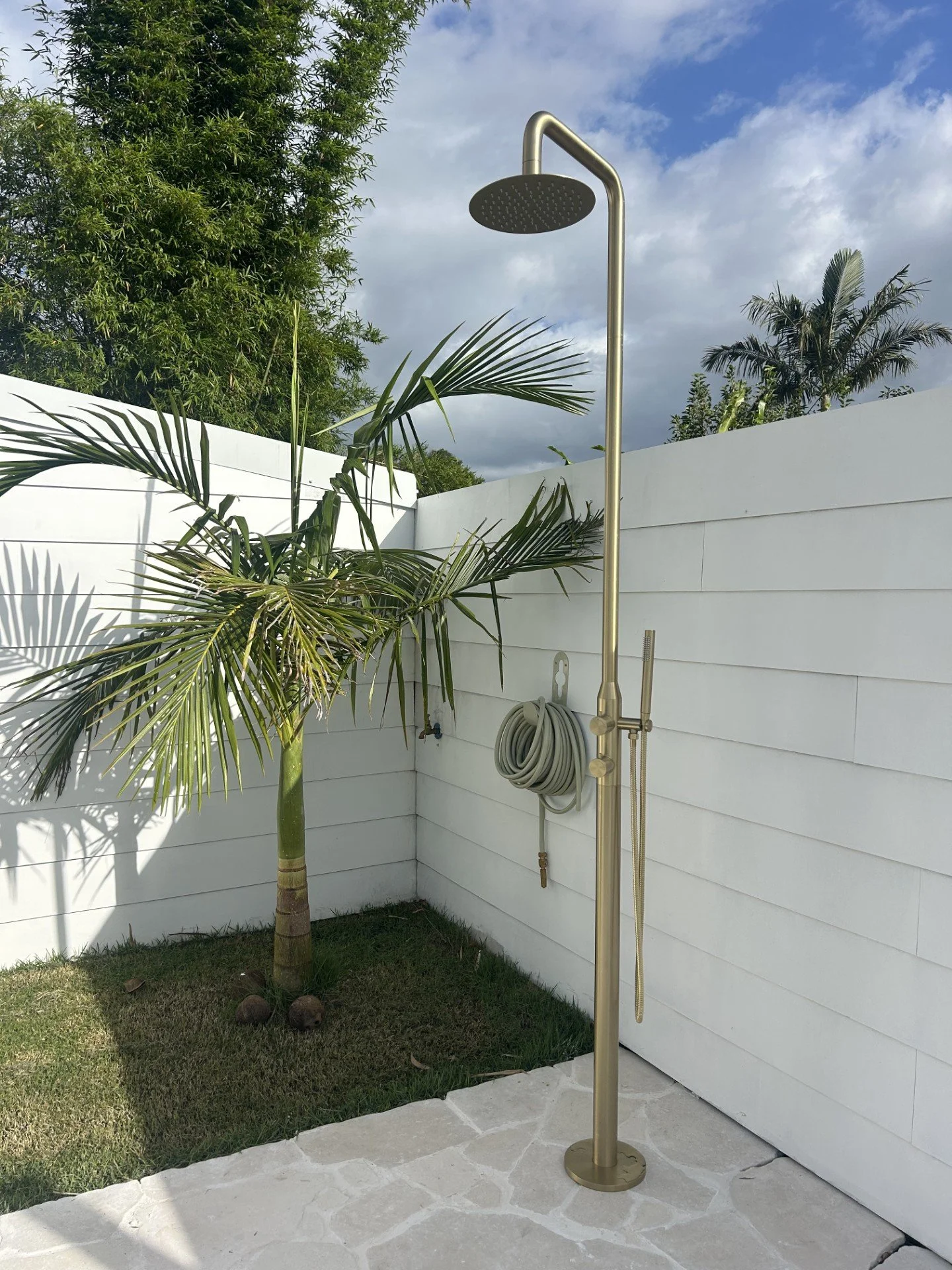Outdoor shower with a large overhead rain shower head, mounted on a pole next to a white wall and a small palm tree, under a partly cloudy sky.