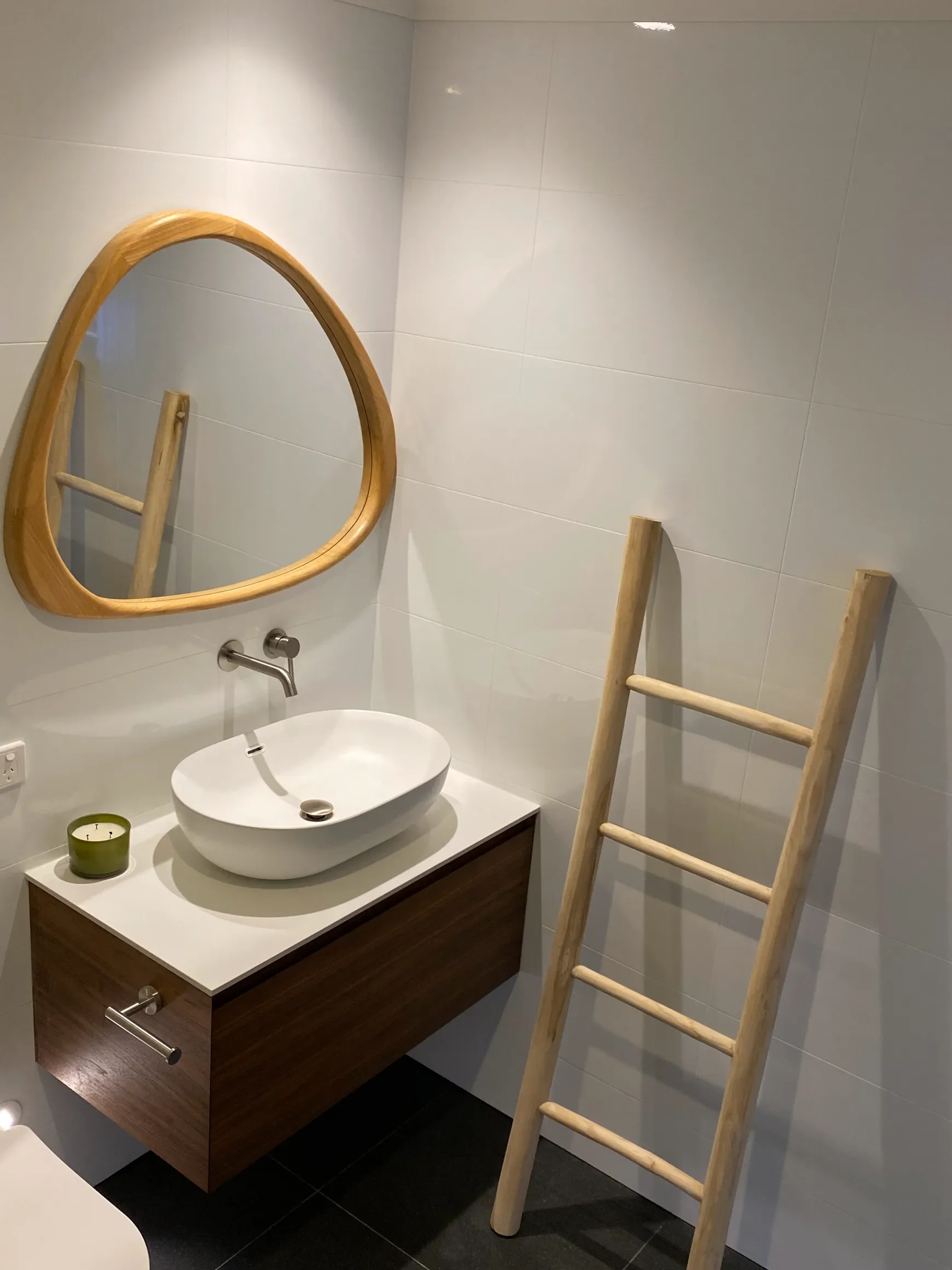 Modern bathroom with a white vessel sink on a wooden vanity, a round wooden-framed mirror, a small green candle, and a wooden ladder leaning against the tiled wall.