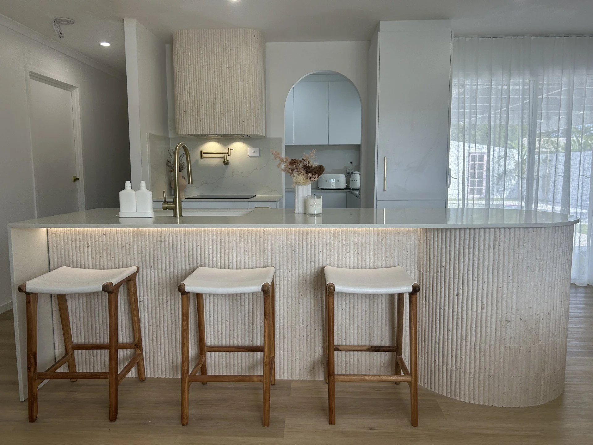 Modern kitchen renovation with a curved island bench, with marble tiles , three wooden stools with white cushions, white cabinetry, a gold tapware faucet, and a large window covered by sheer curtains, strip lighting. Kitchen inspiration. 