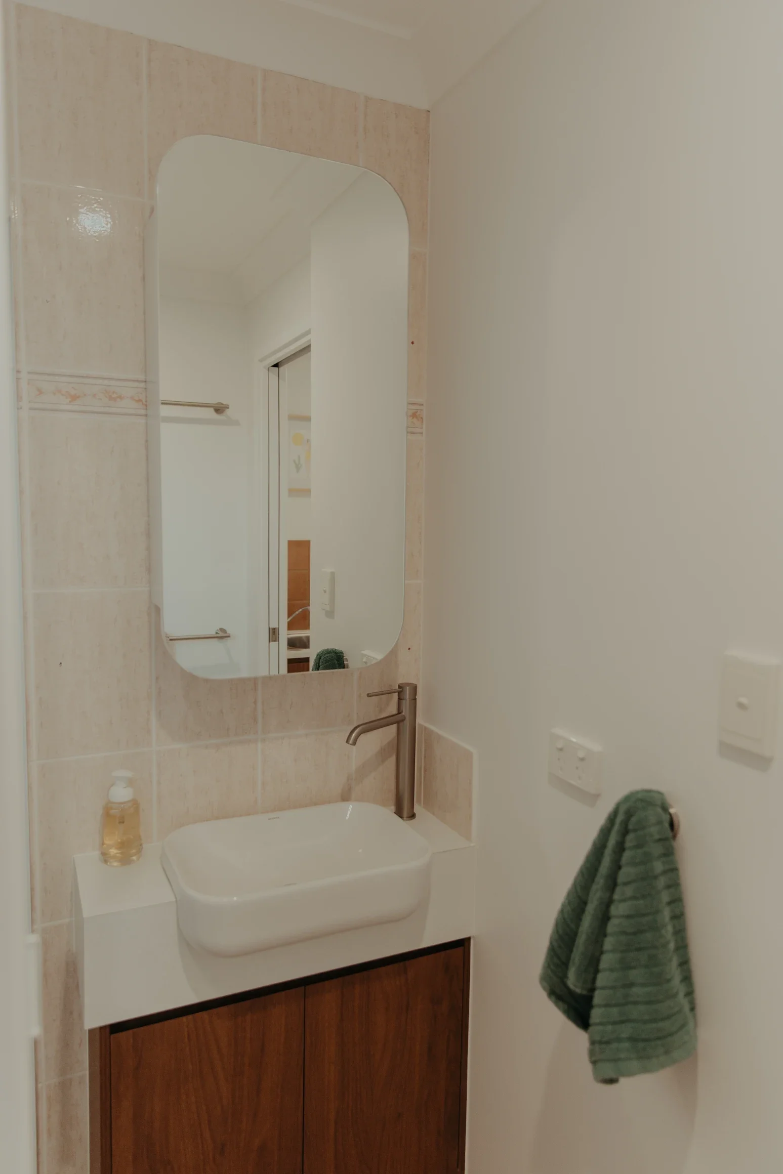 A bathroom vanity with a white sink, gold soap dispenser, and a rectangular mirror above it. To the right, a green towel hangs on a white wall with electrical outlets.