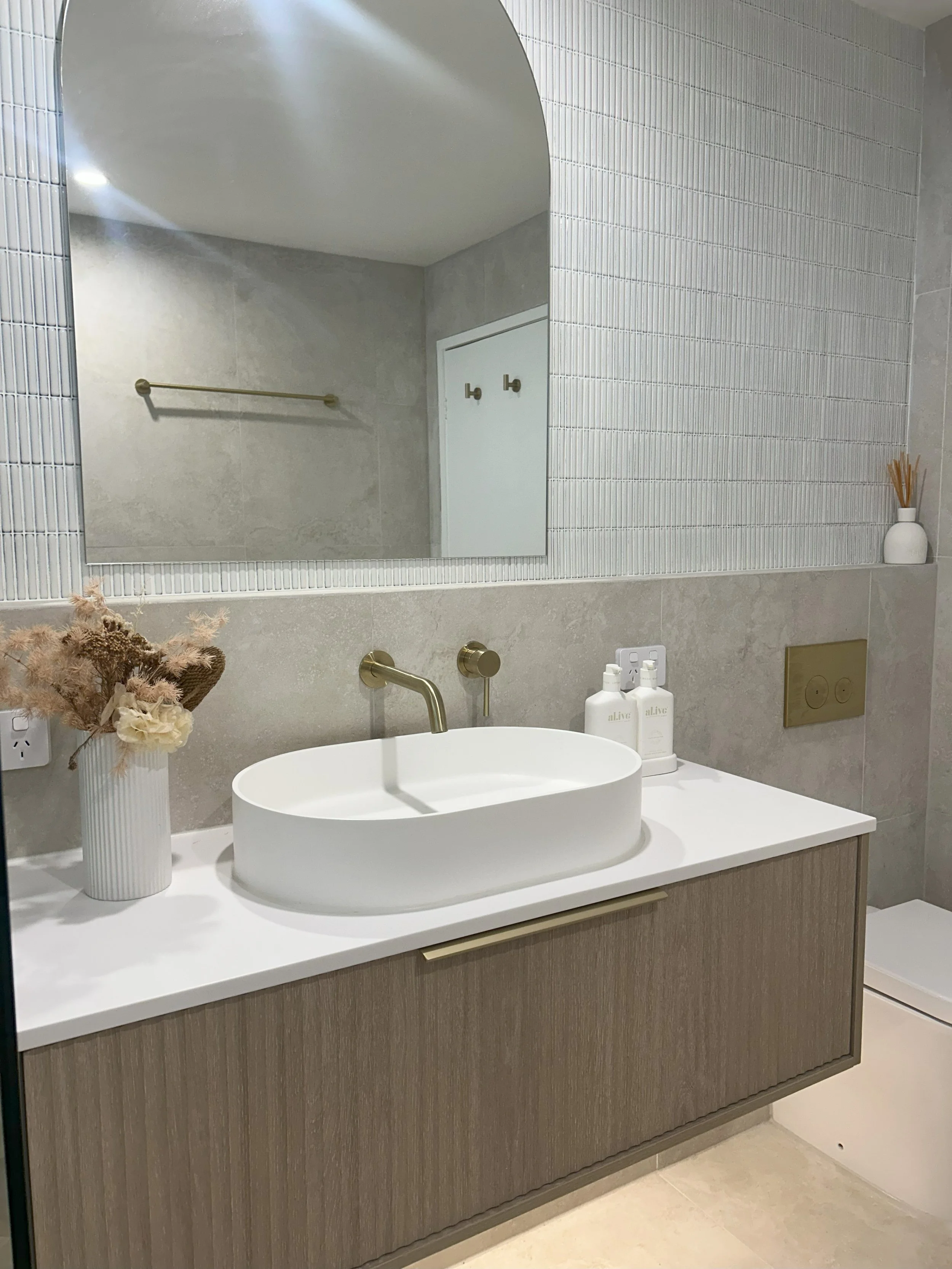 Modern bathroom vanity with a round vessel sink, gold fixtures, and minimal decor, including a vase with dried flowers and a white bottle set.