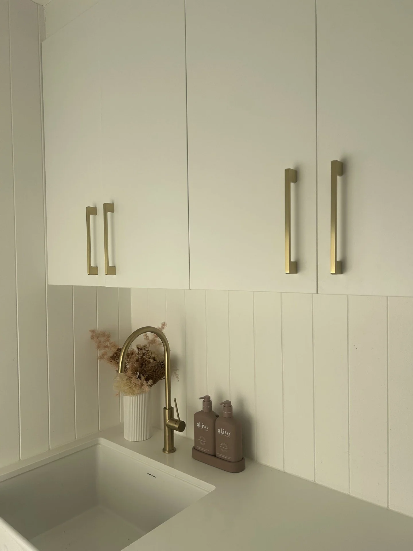 Kitchen sink with a gold faucet, beige wall cabinets with gold handles, a white countertop, a decorative vase with dried flowers, and two bottles of hand soap.
