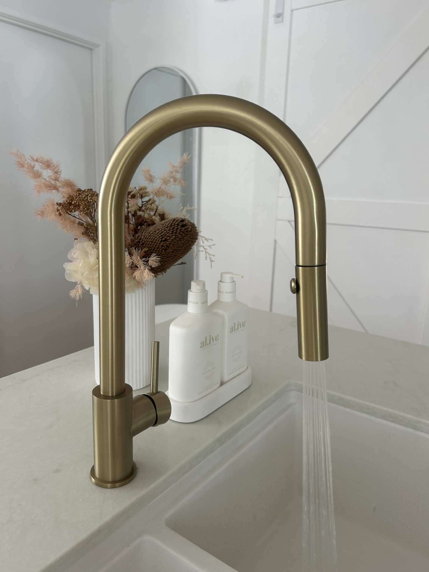 A modern kitchen sink with a matte gold faucet, running water, a white countertop, a ceramic vase with dried flowers, and two bottles of lotion or soap.