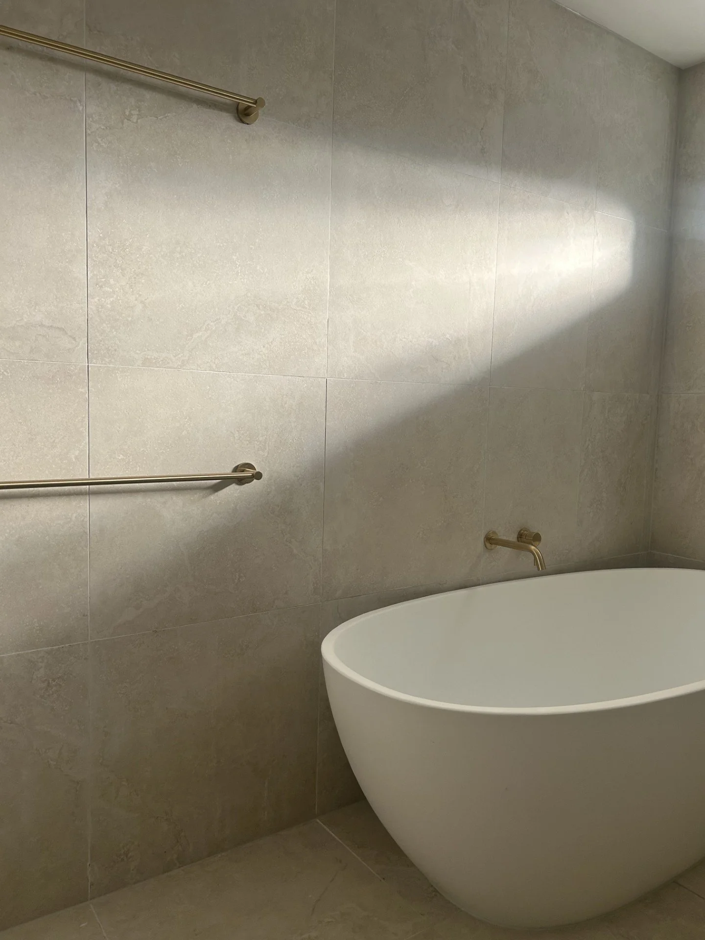A bathroom with beige tiled walls and floor, a white freestanding bathtub, and gold-colored towel bars and faucet. National Tiles, bathroom renovation