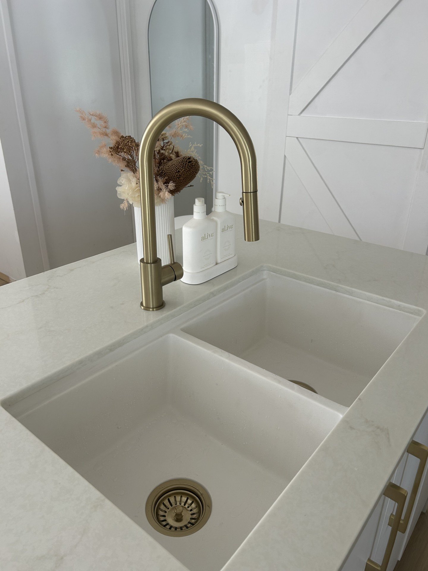 Kitchen renovation, inspiration. A white kitchen sink with a brushed gold faucet tapware and running water, an Alive white soap and lotion dispenser, and a vase with dried flowers in the background. Sunshine Coast, Noosa, Brisbane