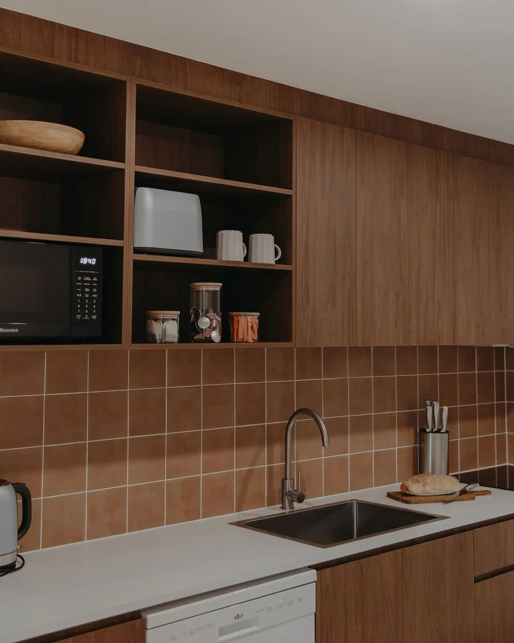 Kitchen with brown warm wooden cabinets, brown tiled backsplash, stainless steel sink, brushed nickel tapware, microwave, toaster, and countertop with bread and utensils. kitchen renovation on the sunshine coast. 