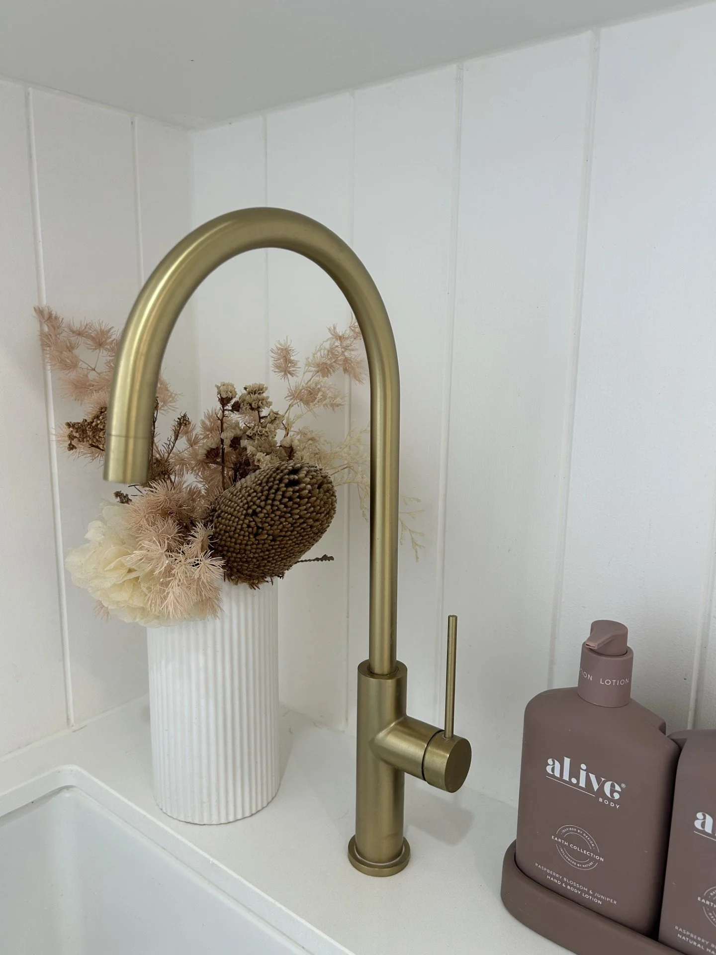 A kitchen or bathroom sink area with a gold-colored faucet. There is a white vase with dried flowers and two bottles of lotion labeled 'al.ive' on the counter.
