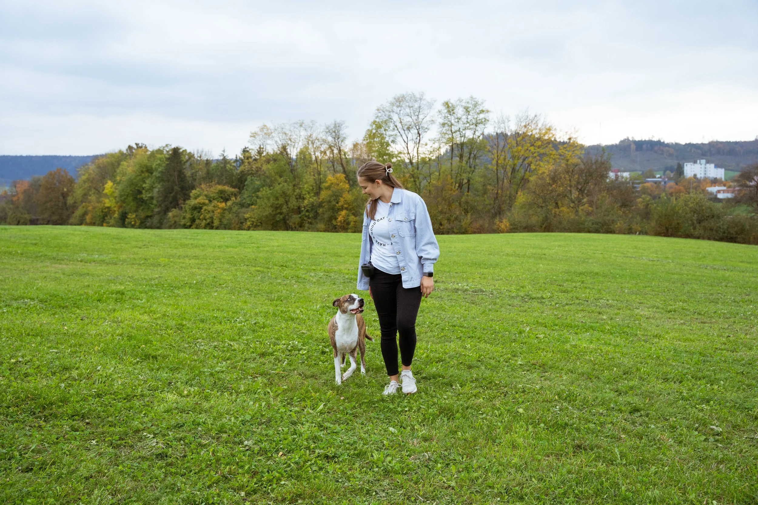 Eine junge Frau mit einem Hund spazieren im Grünen auf einer Wiese an einem bewölkten Tag.