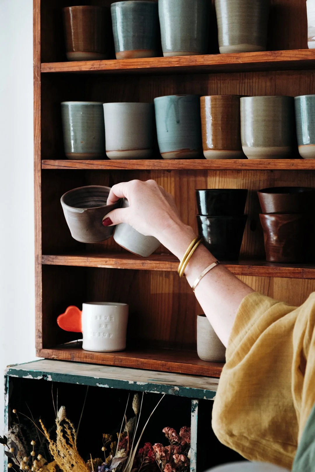 Main d'une céramiste rangeant une tasse en grès sur une étagère en bois remplie de poteries, banque d'images complète