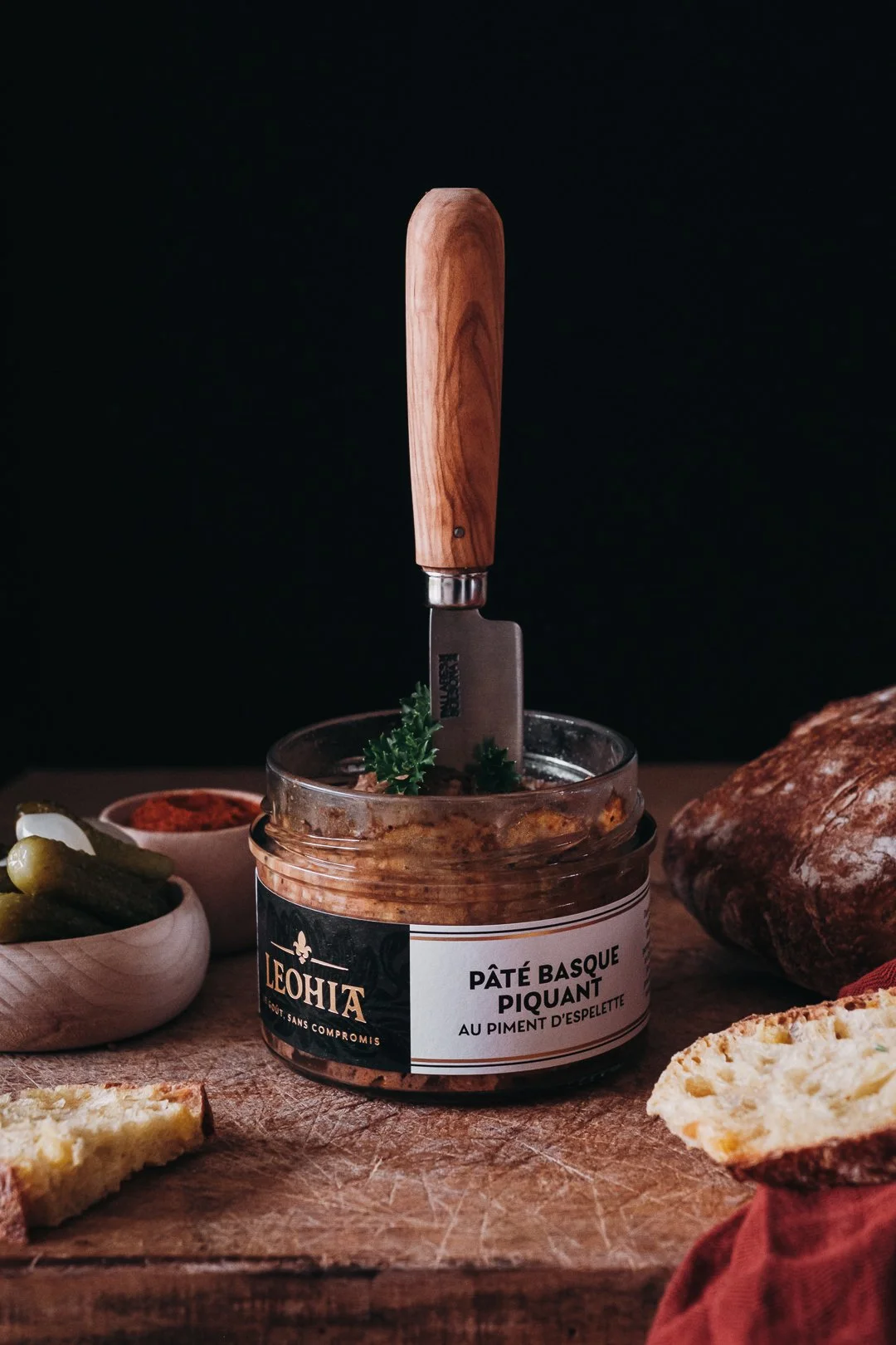 Bocal de Pâté Basque piquant Leohia avec couteau planté dedans, entouré de cornichons et piment, identité visuelle food.
