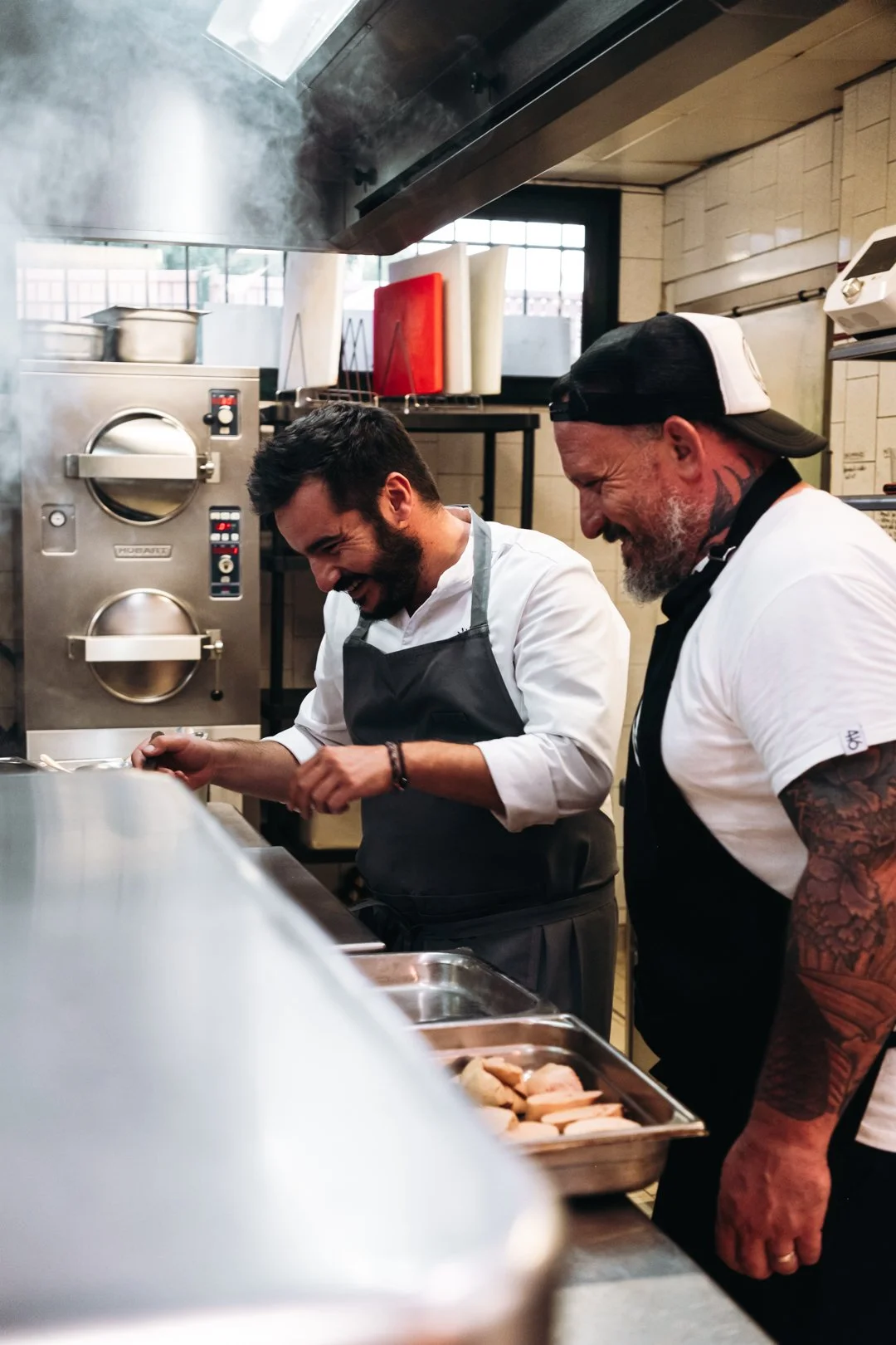 Deux cuisiniers riant devant un four en inox avec de la vapeur, ambiance de service chez Maynats.