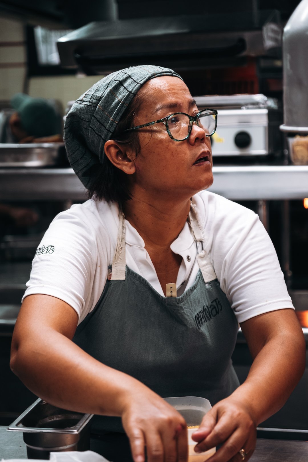 Portrait d'une cuisiniere en tablier Maynats, regard concentre pendant le service.