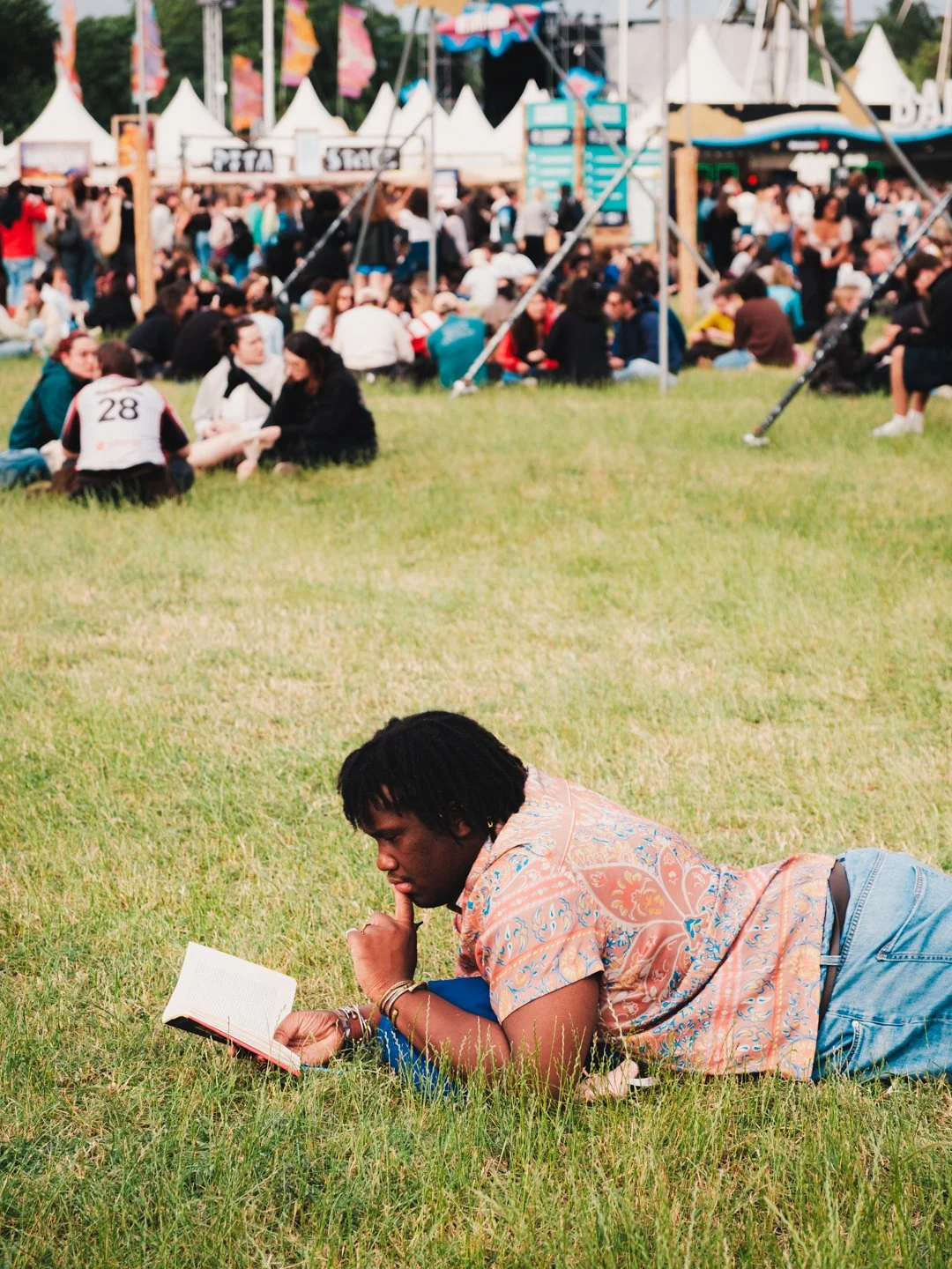 Festivalier allonge dans l'herbe lisant un livre, stands et drapeaux en fond, We Love Green.